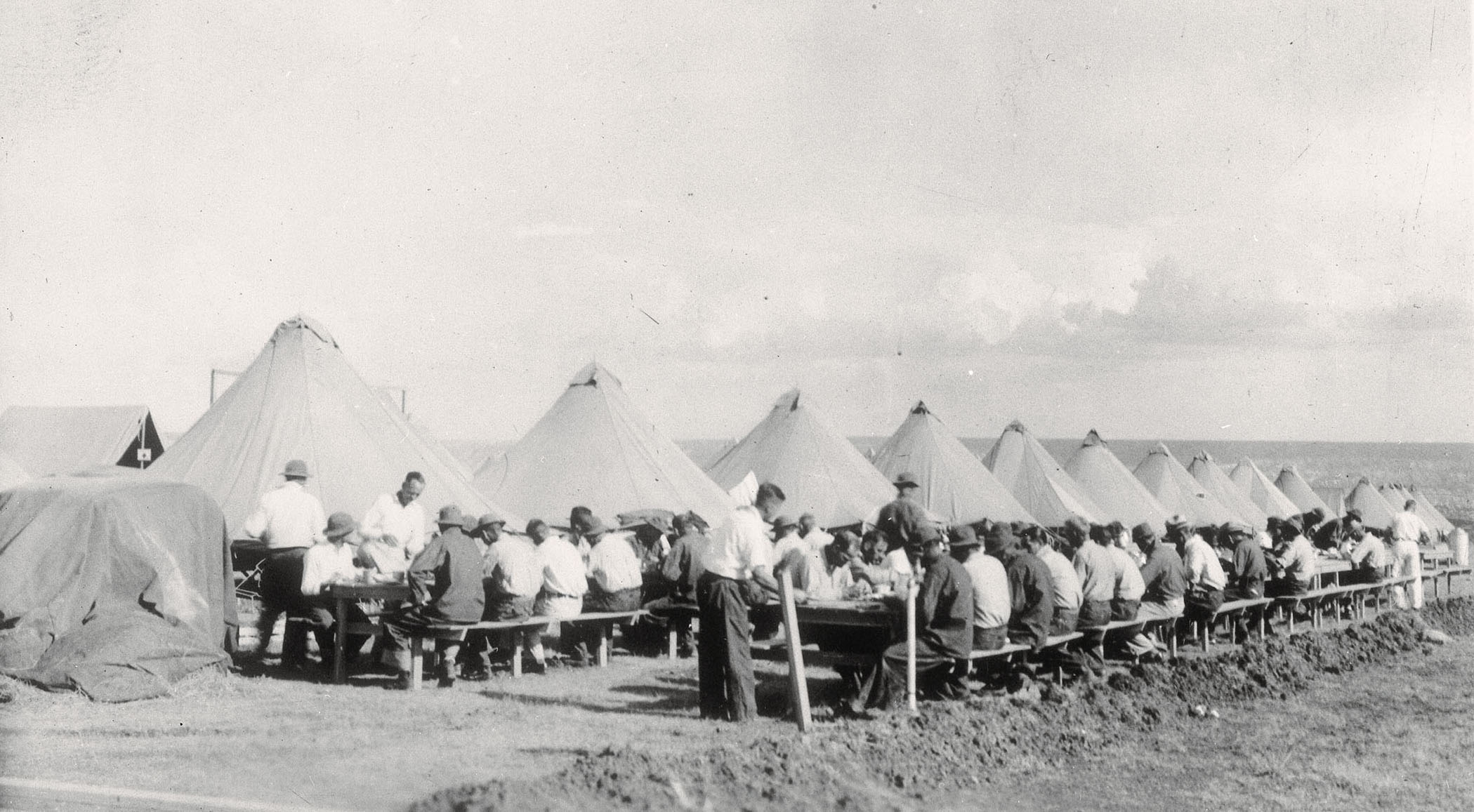 A group of people in white shirts sit at long tables next to tents outdoors