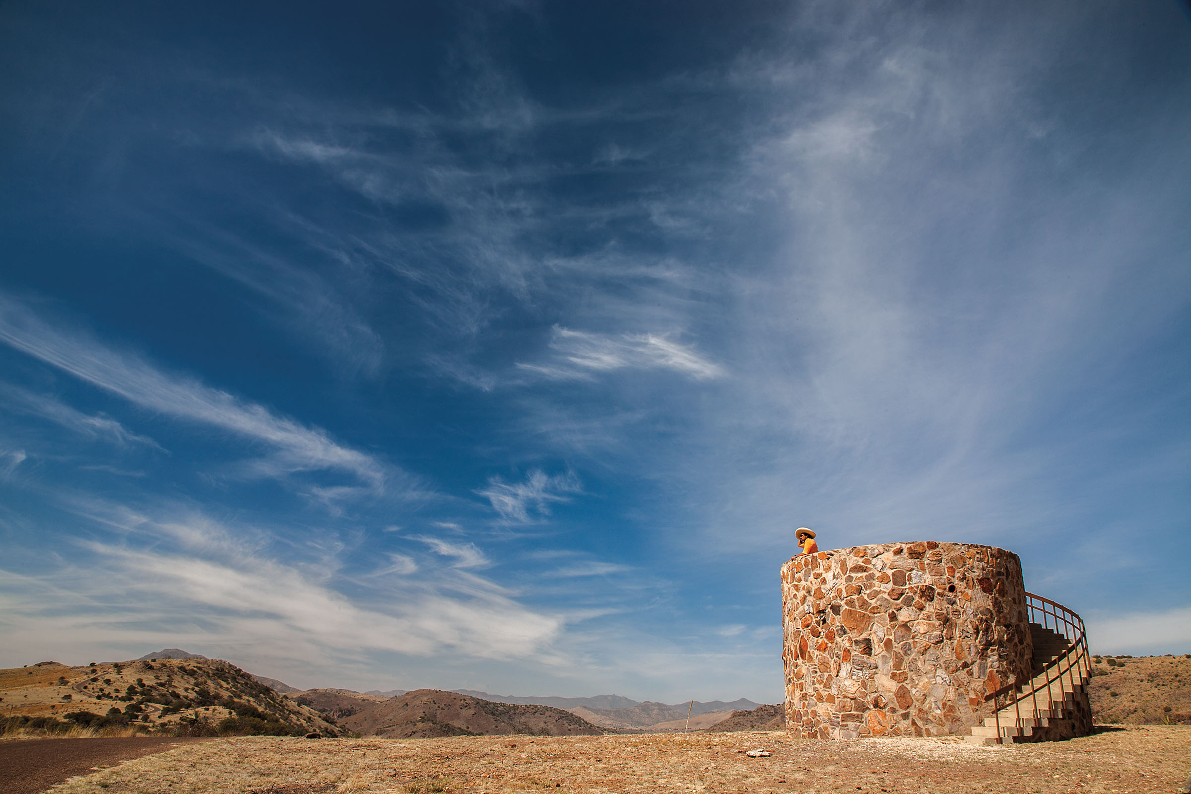 A woman stands on top of a large stone lookout tower under blue sky in the mountains