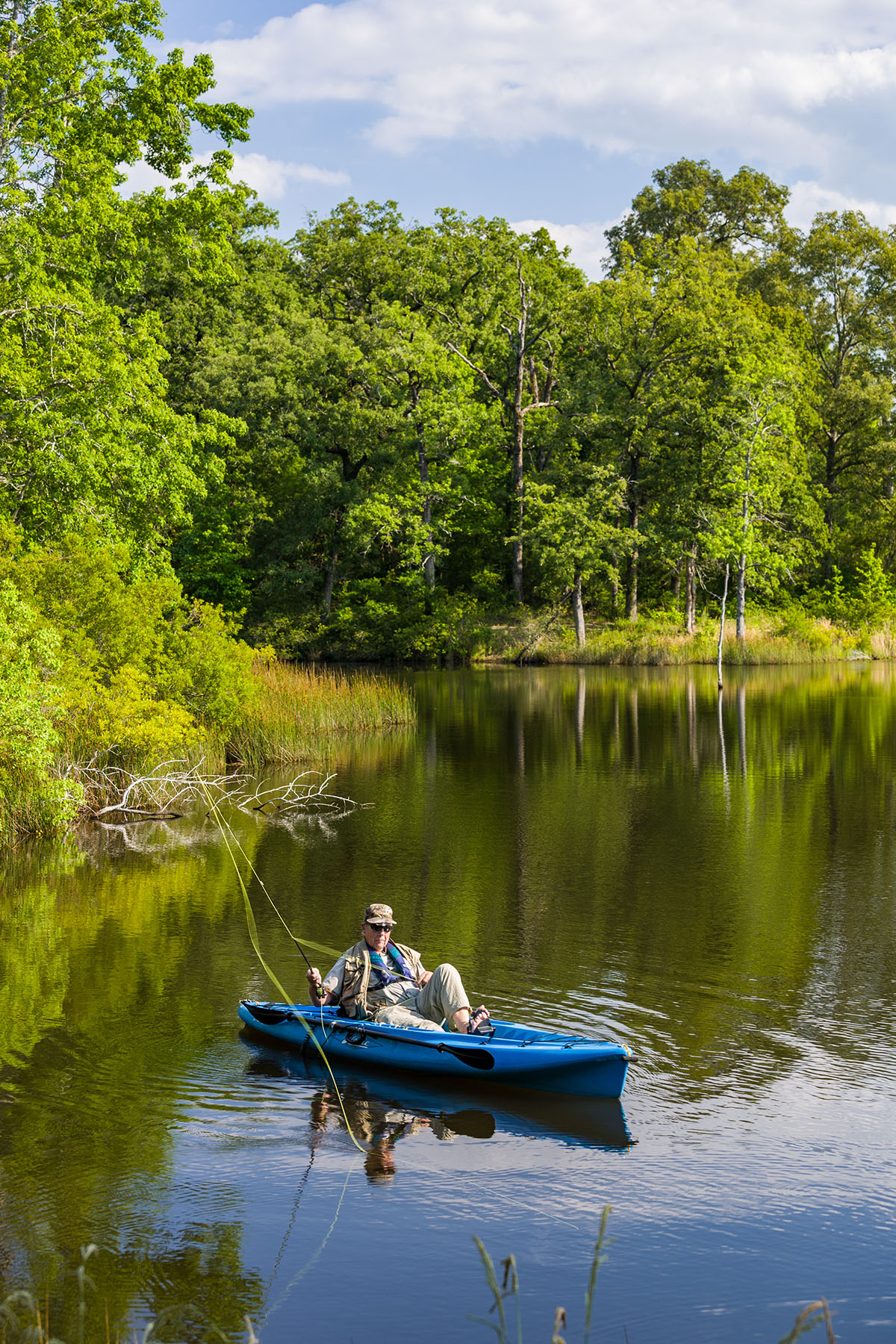 A person in a blue kayak casts a long fishing line on still water in front of blue trees
