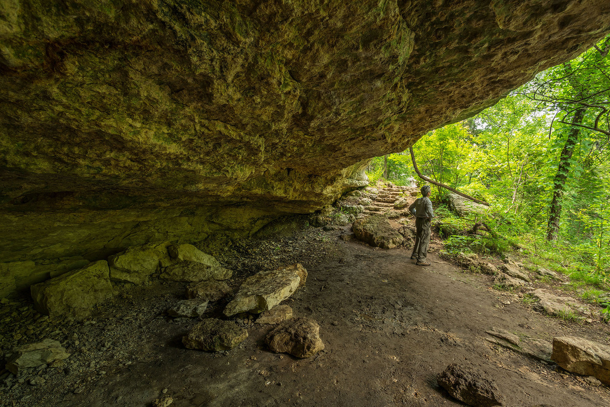 A hiker stands under a large rock outcropping surrounded by green trees