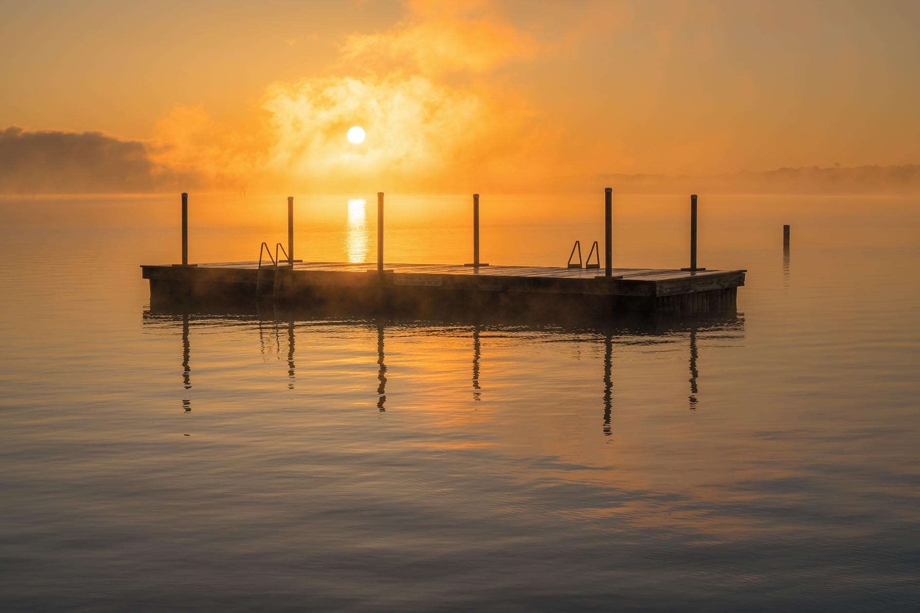 The sun rises over the misty water and a swimming platform out on a lake.