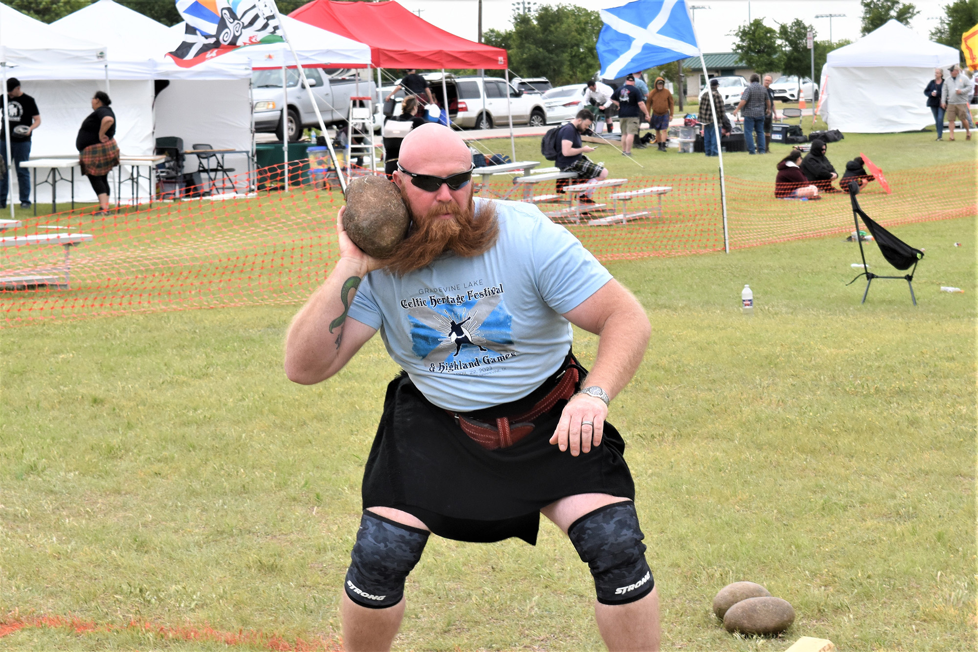 A man with a beard wearing a Celtic Heritage Festival T-shirt and kilt holds a shot put against his neck to prepare for a throw
