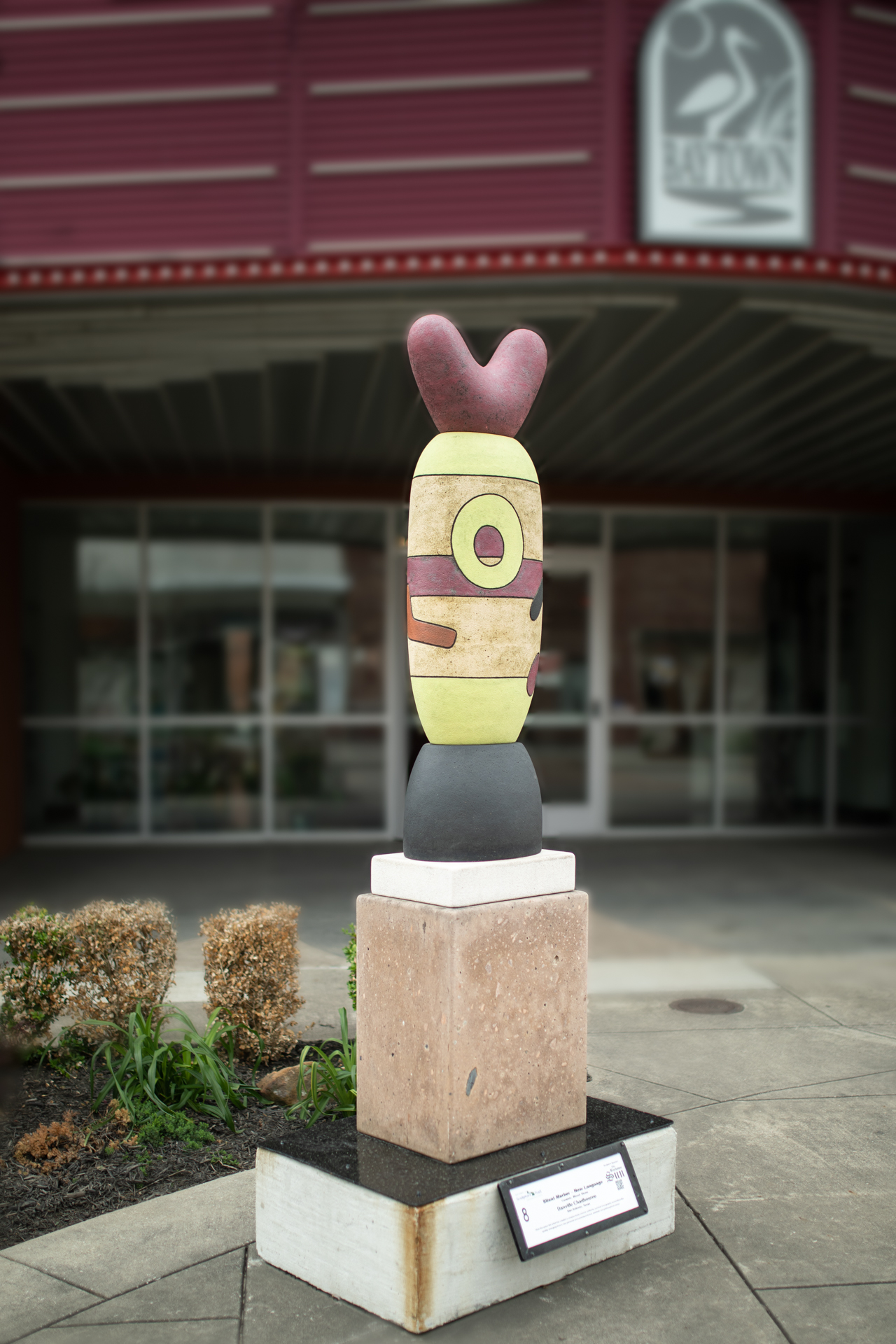 A small yellow sculpture with an eye-like design and red top in front of a building