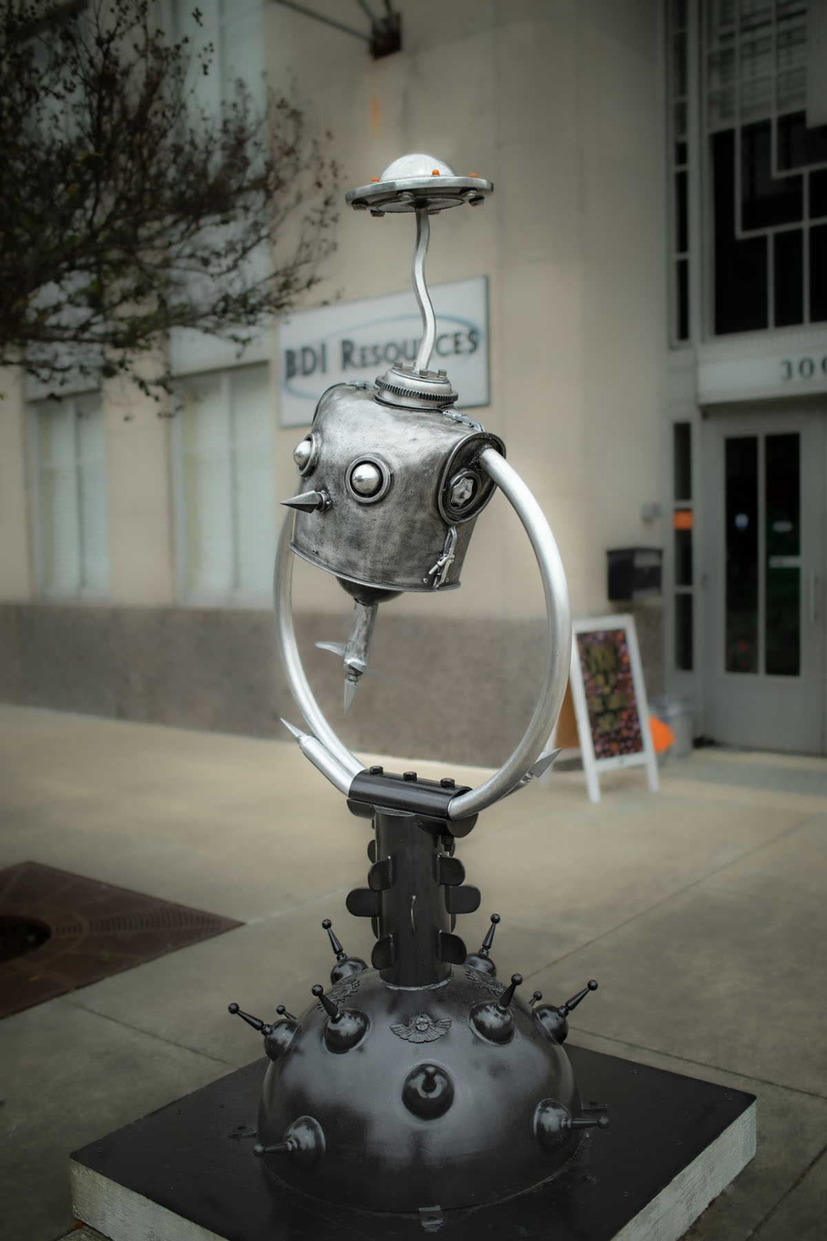 A metal sculpture with a face-like top and steel arms
