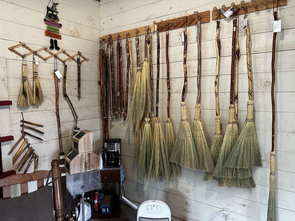A variety of brooms styled like those of the 1830s hang from a wooden peg rack on the wall of the store. 