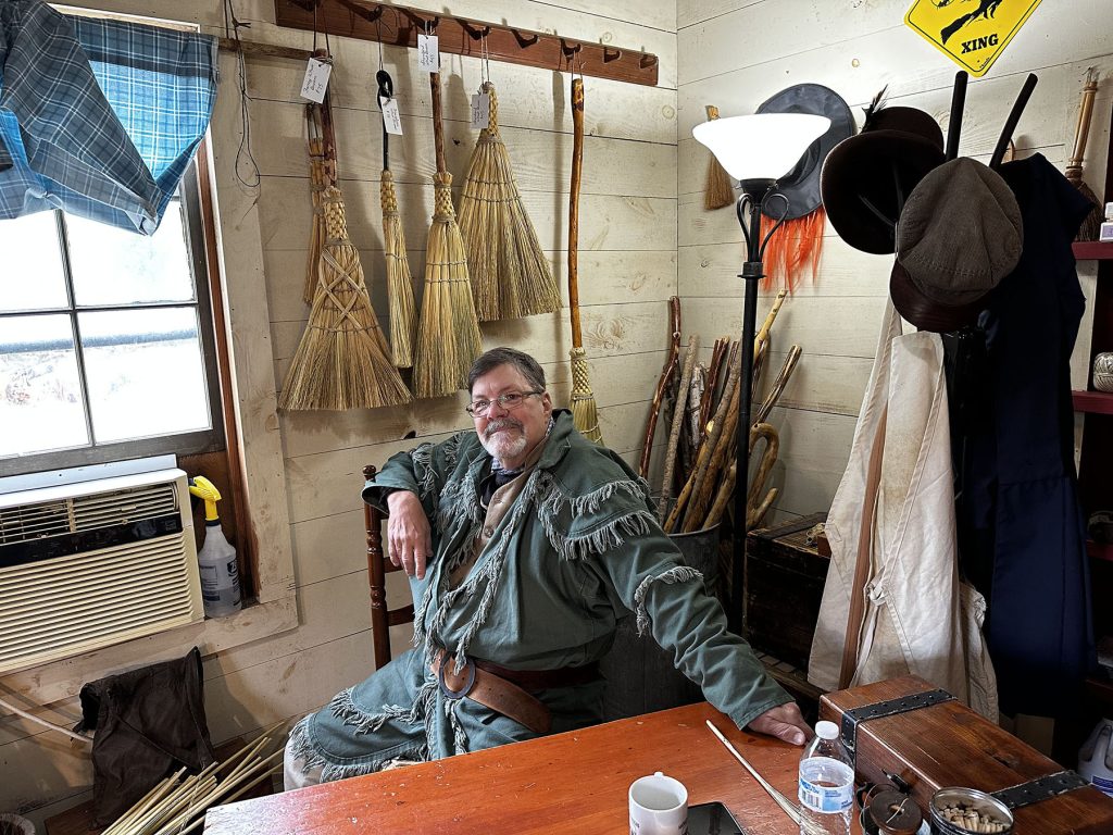 John Potter sits at a desk in the store. He wears glasses and wears a green tunic with fringe and leather belt. Brooms hang on a wooden peg rack behind him. 
