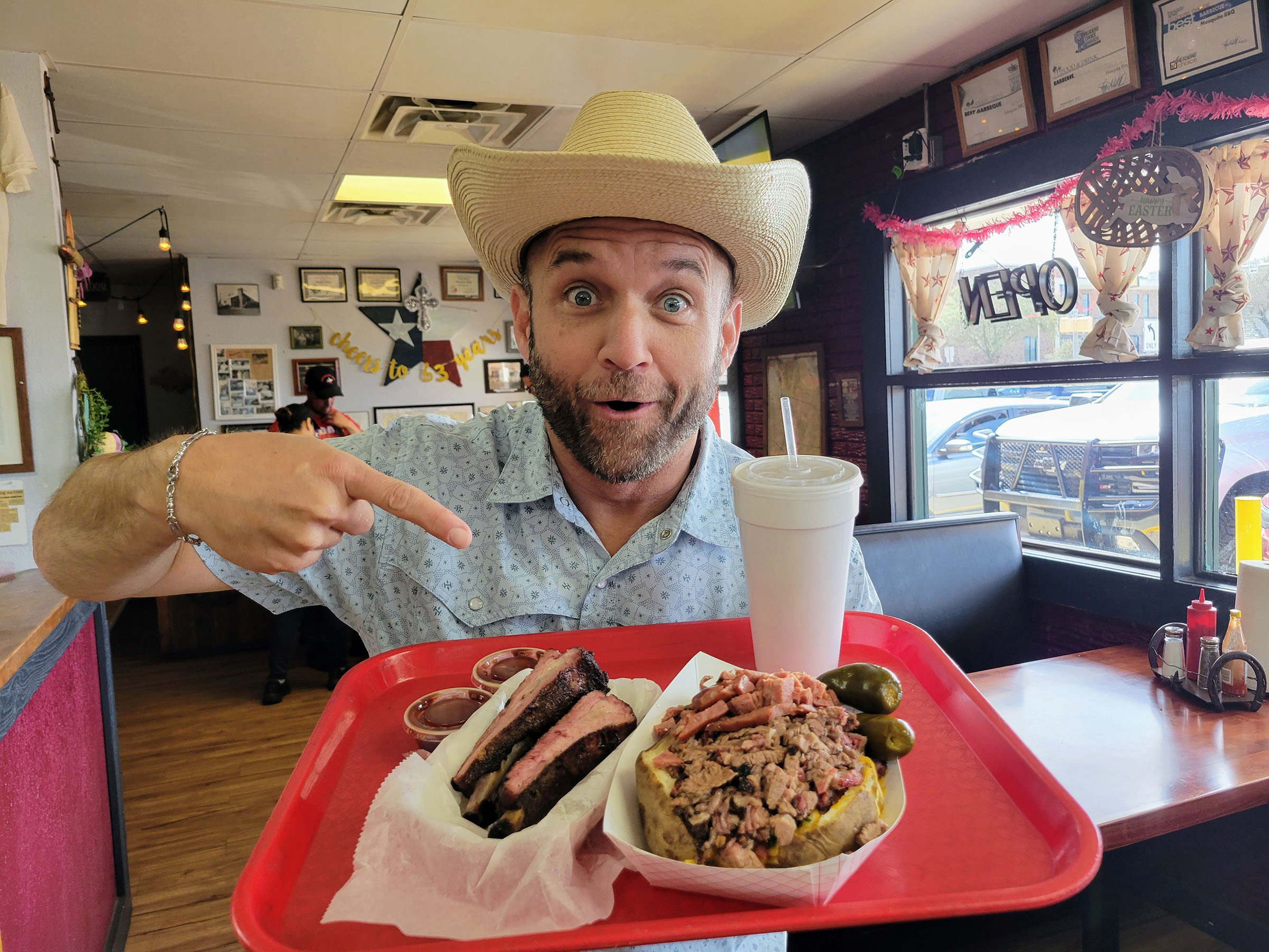 A man in a cowboy hat makes a silly goofy face and points at an assortment of barbecue on a red tray