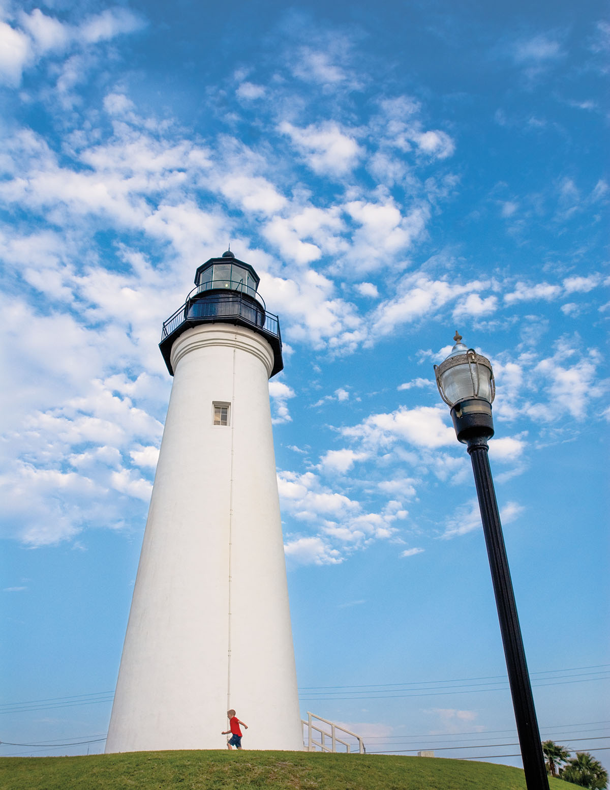 A tall white lighthouse next to a thin black light post in front of a blue sky with scattered clouds