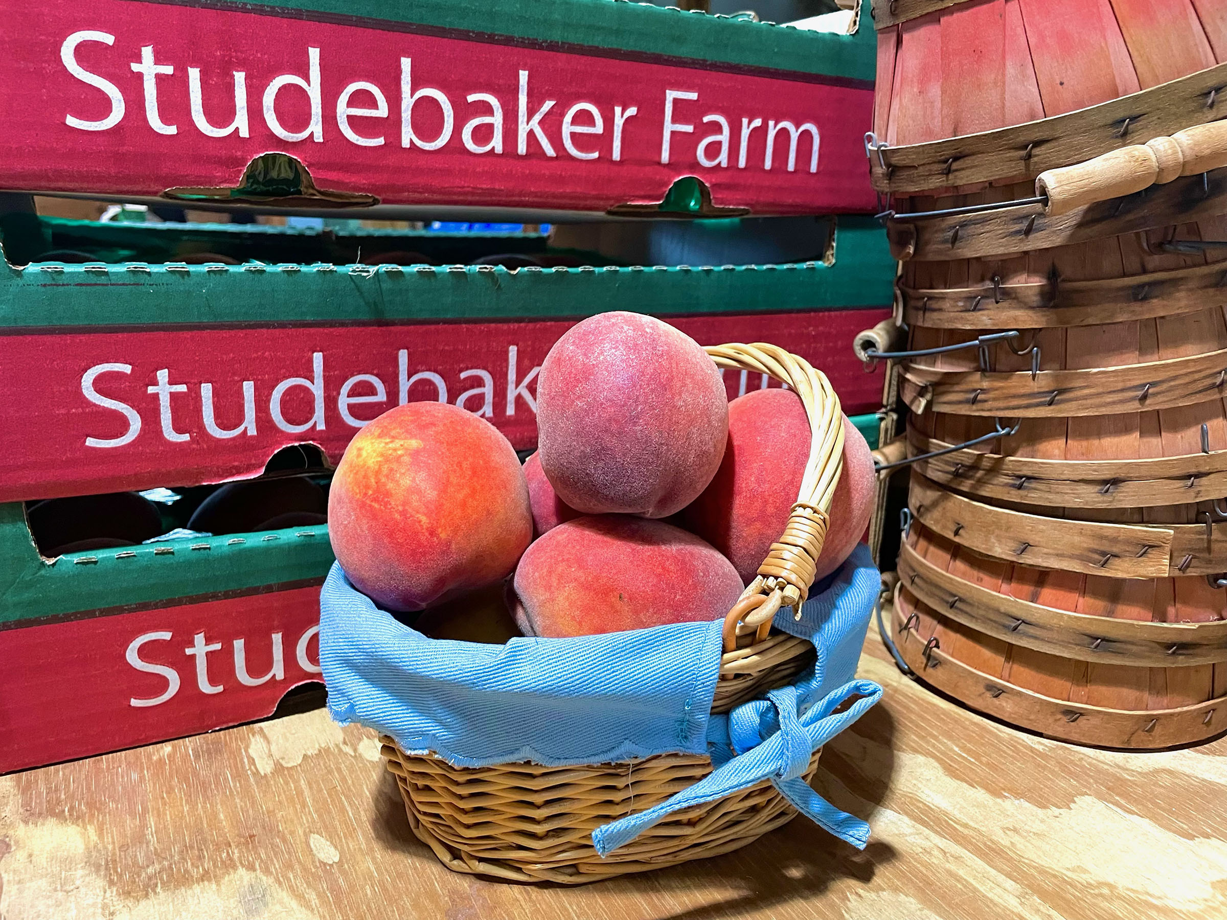 A basket of peaches with a blue napkin in front of boxes reading Studebaker Farm