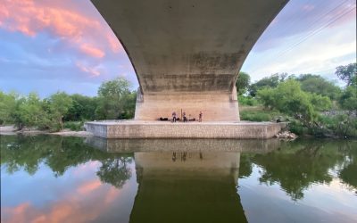 Photo shows underneath the bridge, with a band playing in the distance at the base of an arch, with trees on the sides of the arch. Clouds in the sky are pink from the setting sun.