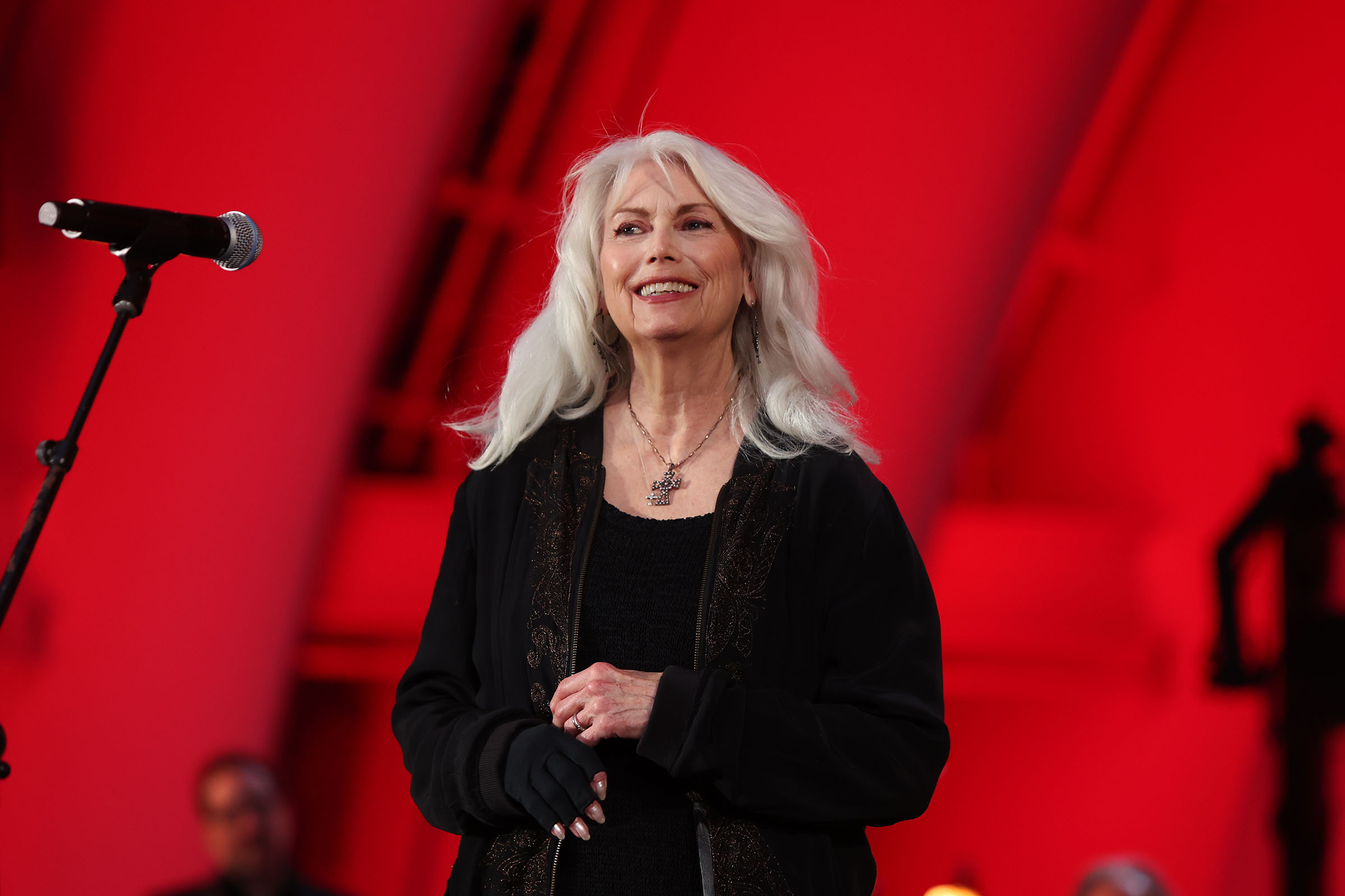 A woman in a black top stands in front of a microphone in front of a red backdrop