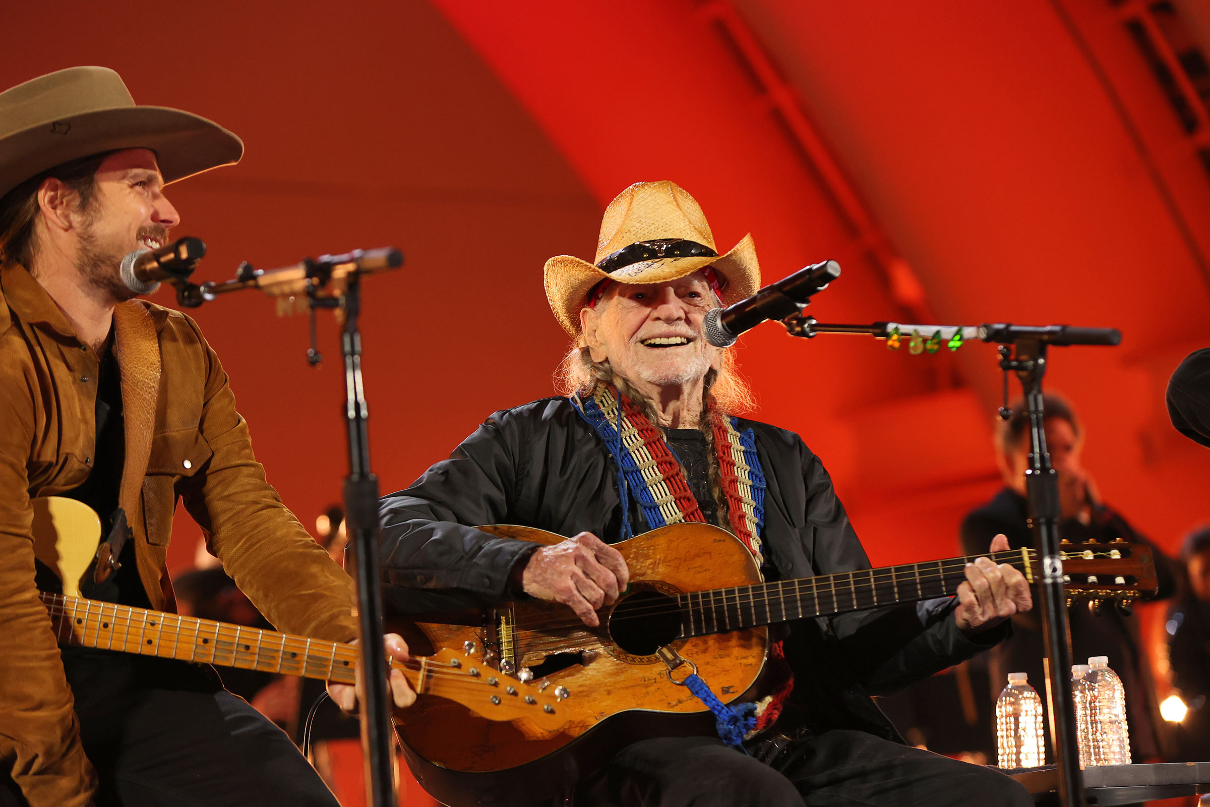 Two musicians holding guitars perform in front of a red curtain backdrop