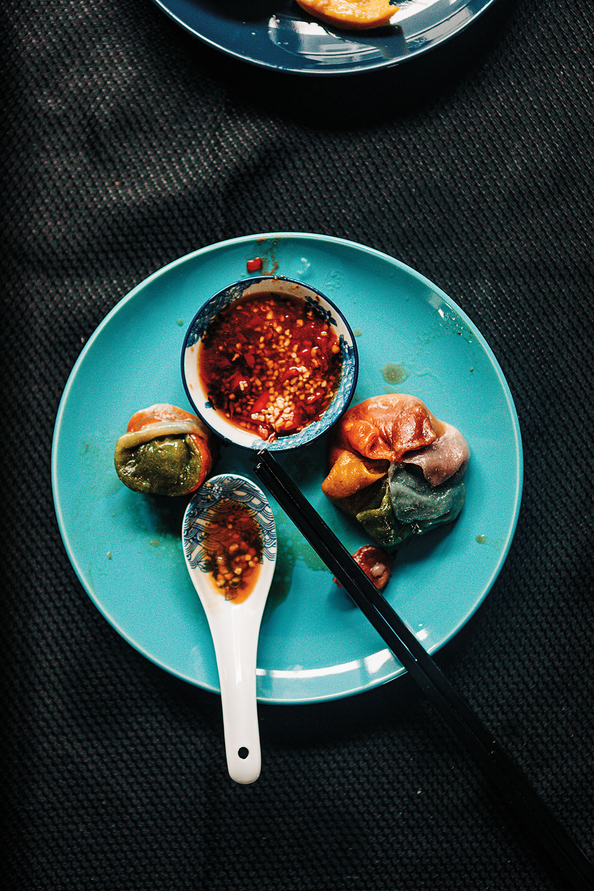 An overhead view of a dumpling, bright red sauce, chopstick and spoon on a teal plate