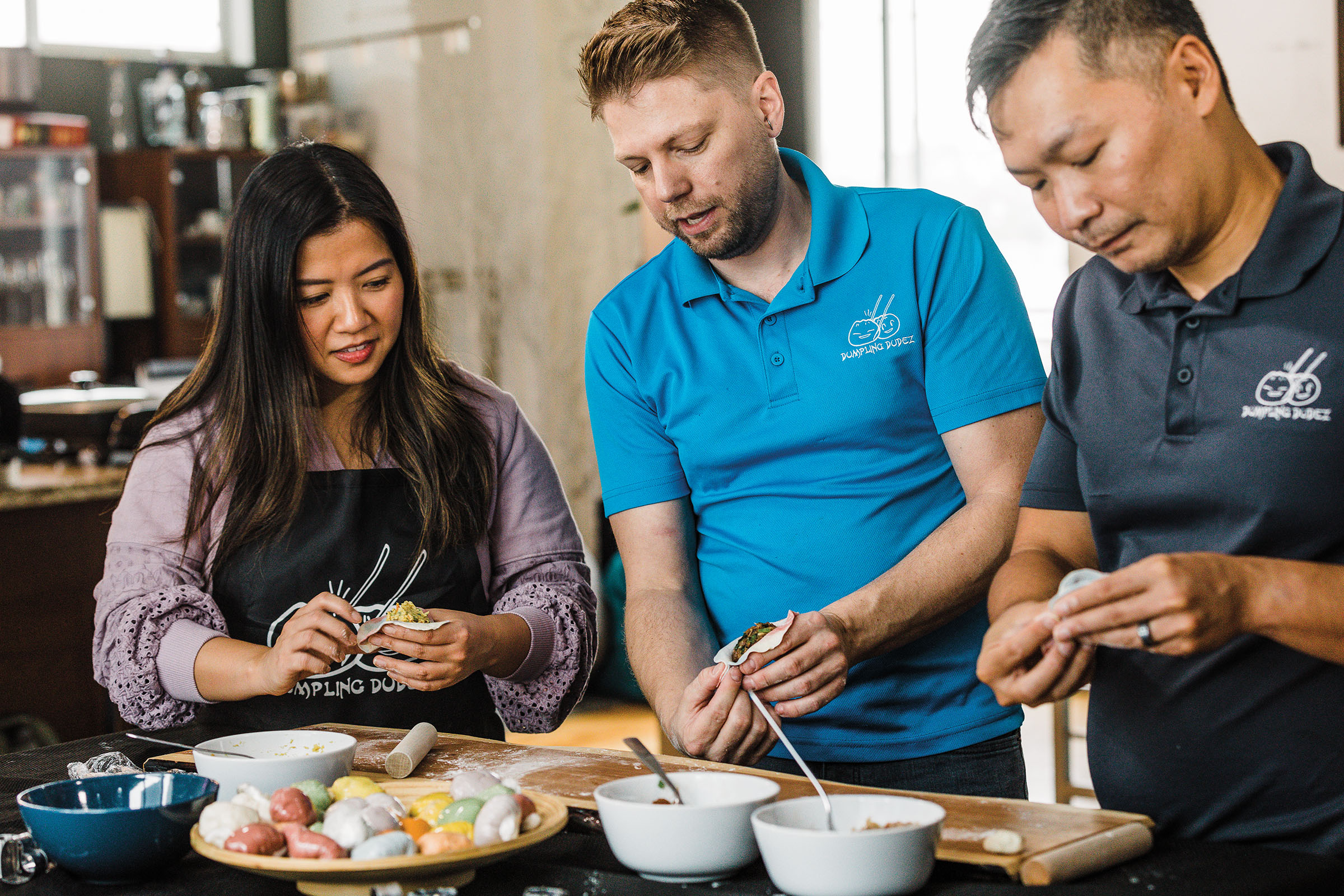 Three people stand at a table carefully filling dumplings from white bowls of various ingredients