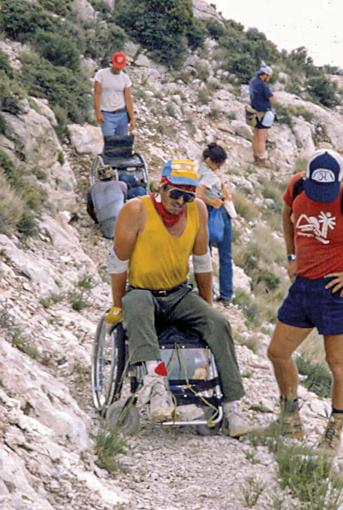 A man in a yellow shirt uses a wheelchair to traverse rocky terrain while several onlookers offer support