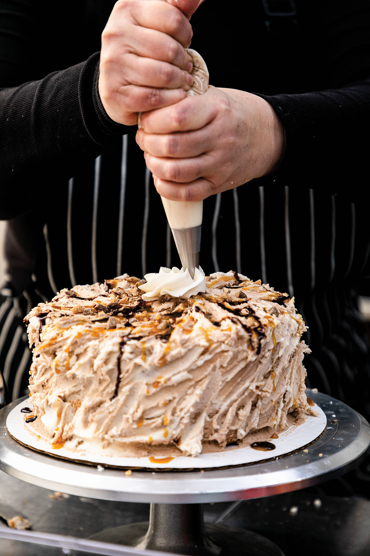 A person carefully pipes white icing onto a chocolate frosted and ganached cake