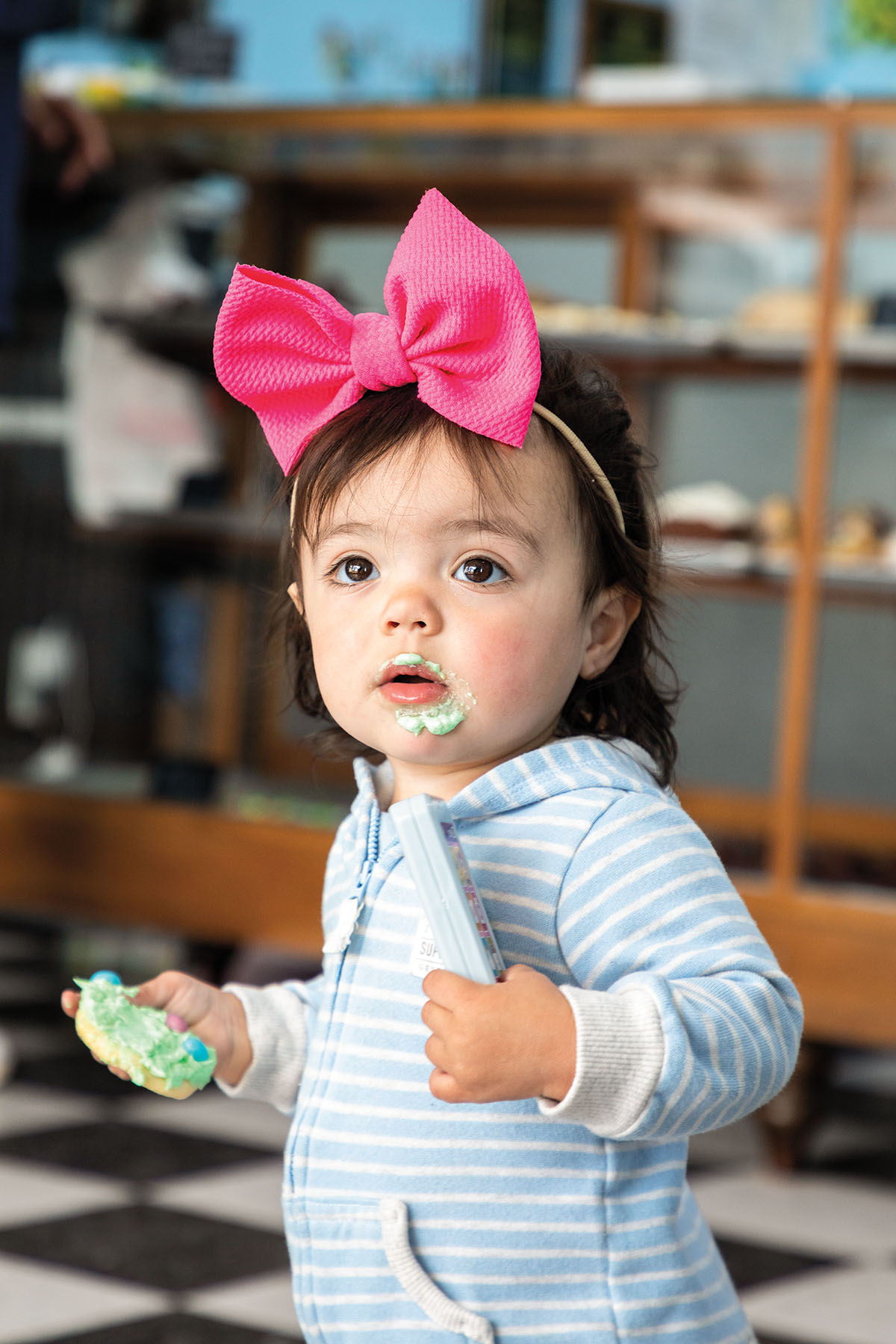 A toddler wearing a bright pink bow holds a cookie in her left hand