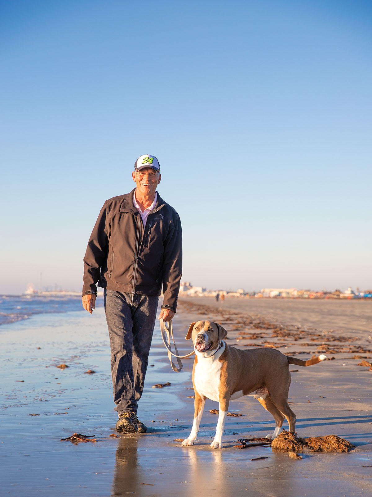 A man in a ball cap walks a dog along a wet sand beach