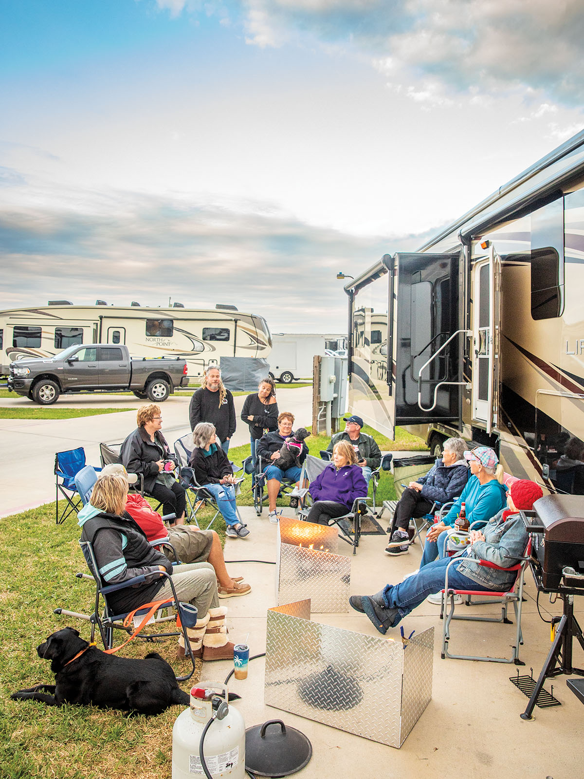 A group of people sit in folding chairs in front of an RV, enjoying drinks and conversation around two gas fire pits