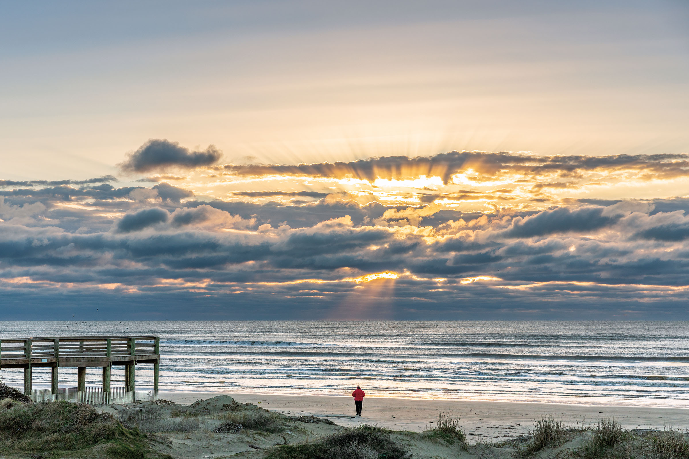A person walks along a beachfront in a morning sunrise with striations of gray clouds and still waves