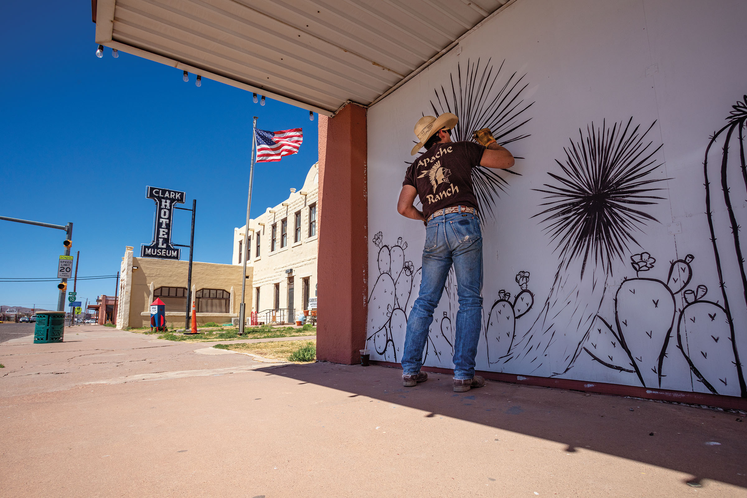 A man in a cowboy hat and blue jeans carefully paints a black and white mural in a desert scene next to an American flag