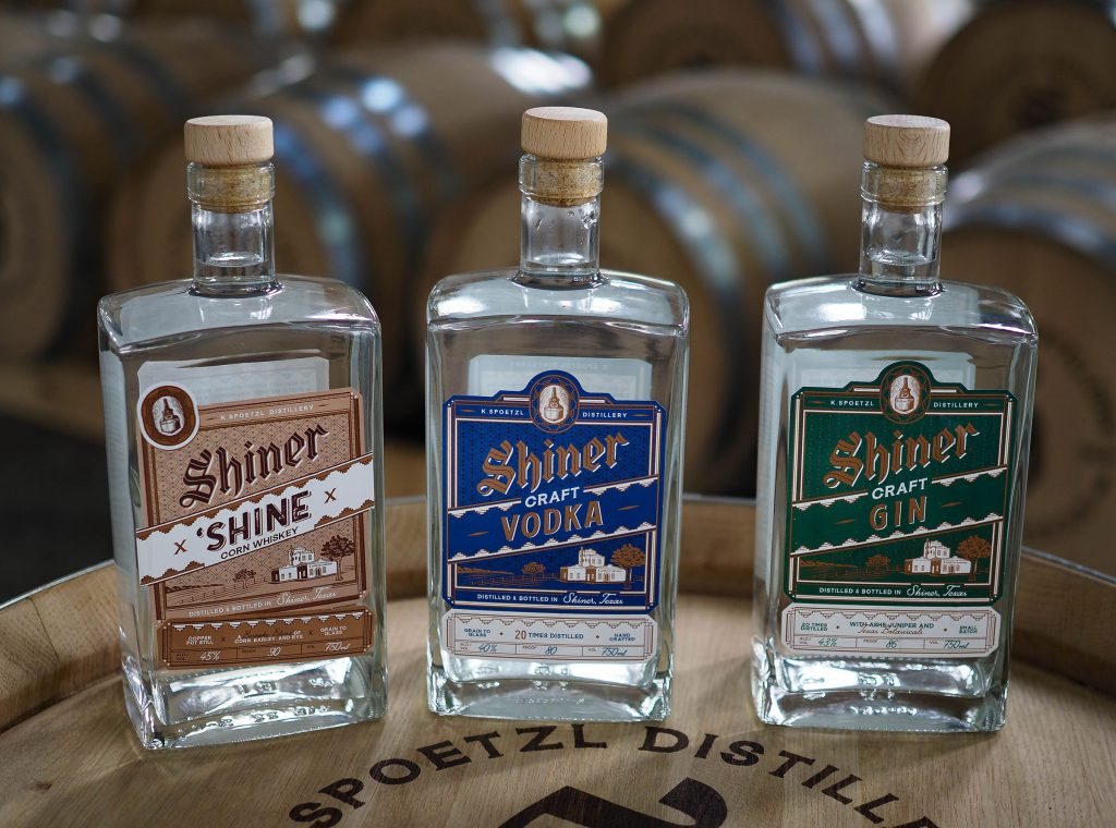 Three rectangular-shaped bottles with corks in them are in the photo. The labels read left to right: Shiner Shine (light brown label), Shiner Craft Vodka (blue label), and Shiner Craft Gin (green label). 