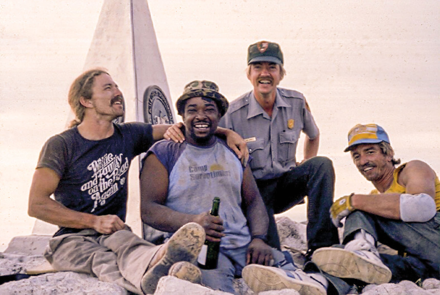 A group of people smile with a bottle of champagne and a park ranger beneath the obelisk marking the summit of Guadalupe Peak