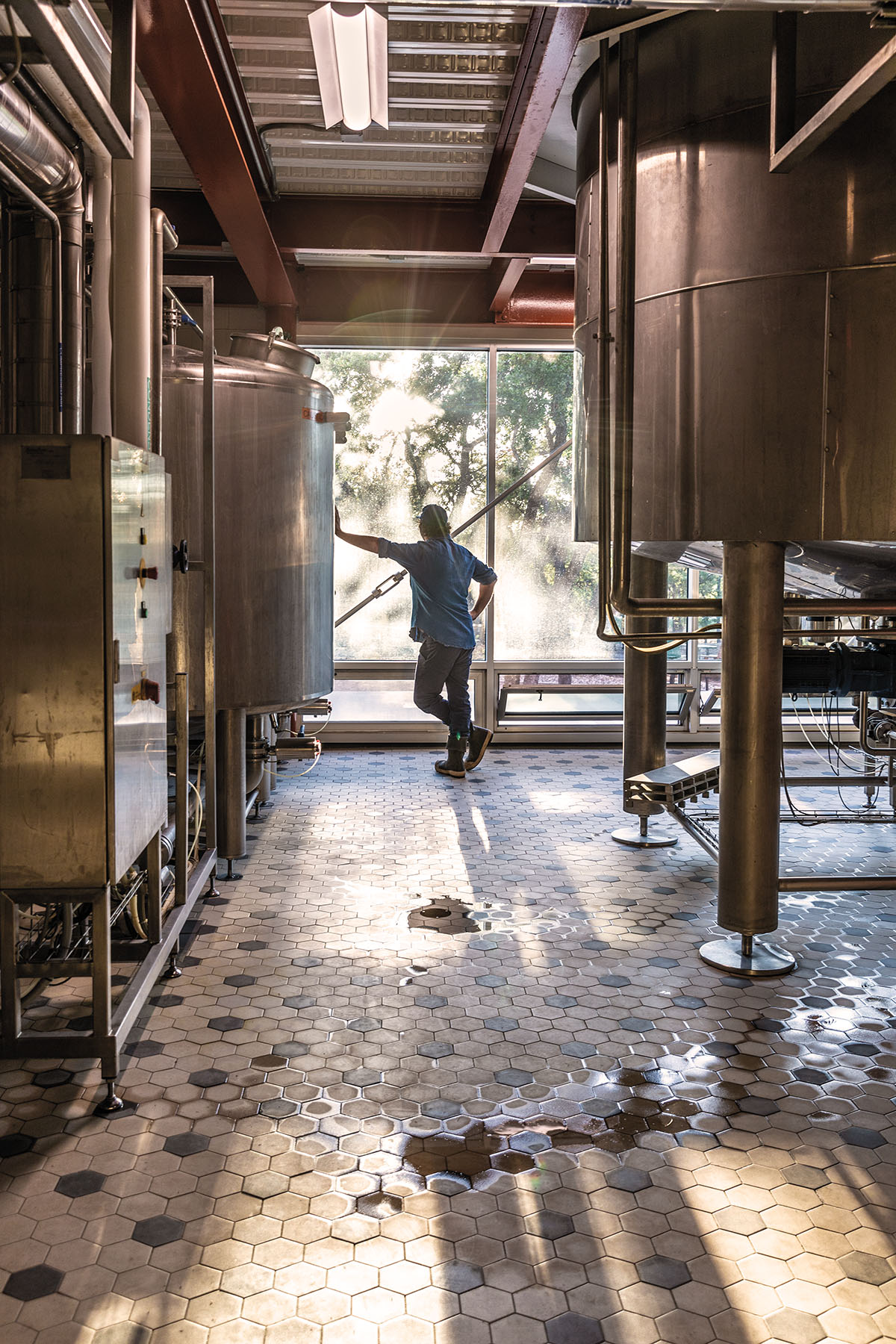 A person stands leaning against a wall behind a large steel brewing tank