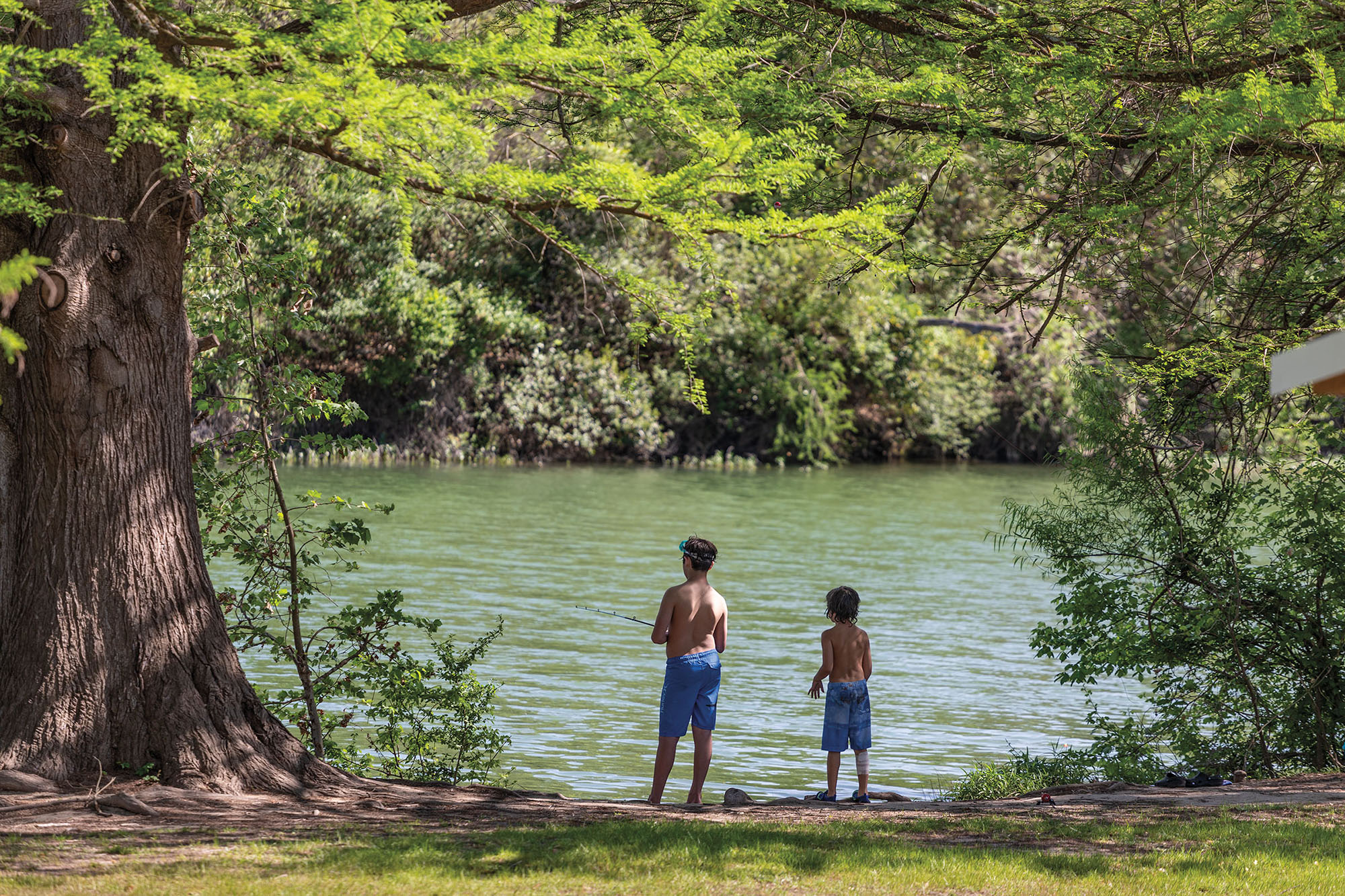 Two young people stand along the banks of a river under green trees