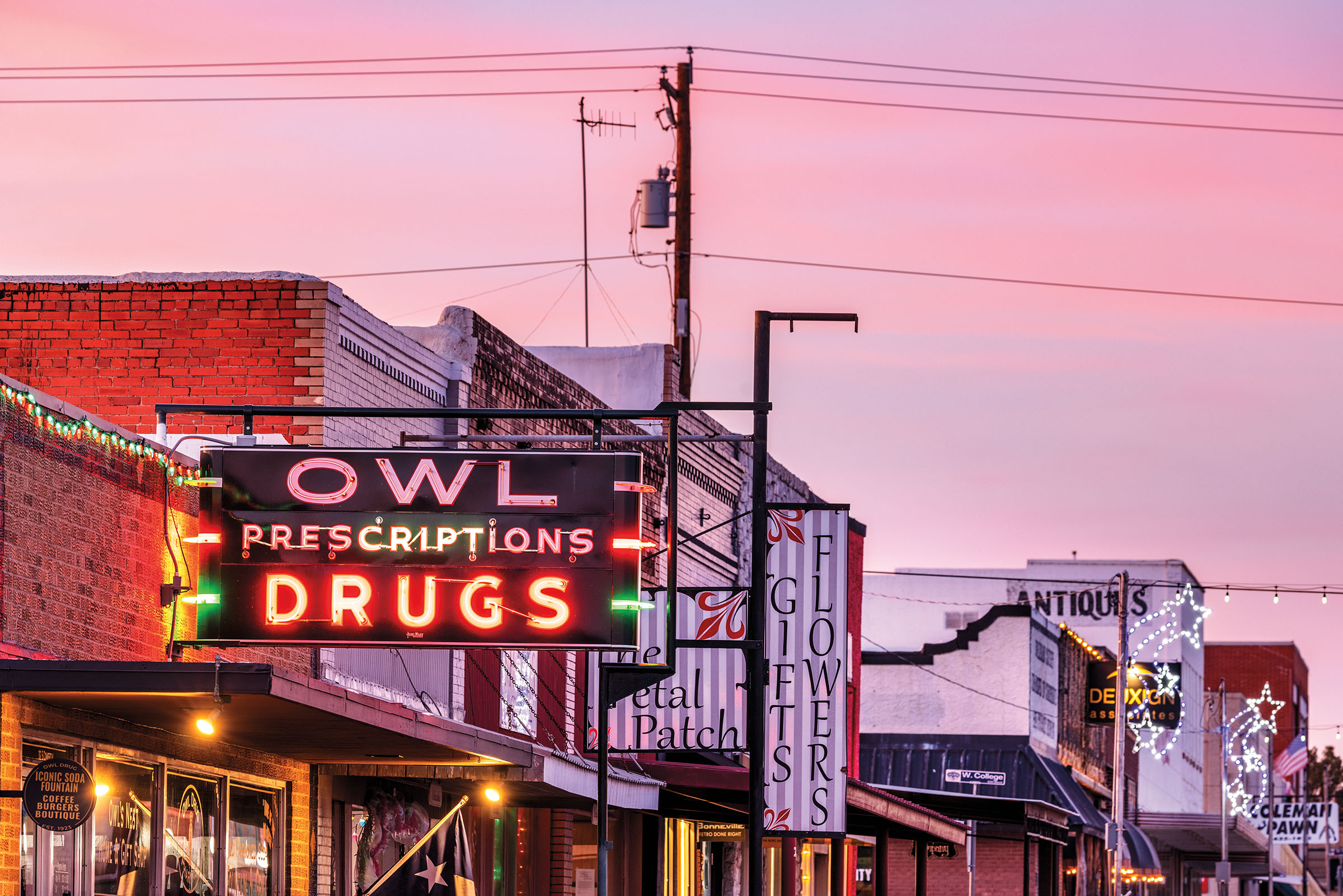 A neon sign reading 'Owl Drug' at sundown on a quiet street