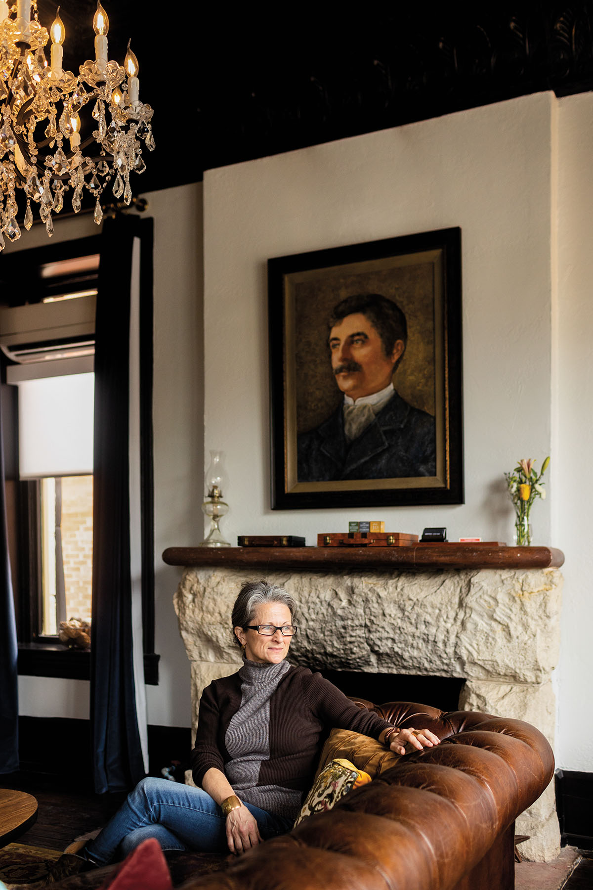 A woman sits on a large leather chair beneath a painted portrait in a richly decorated room