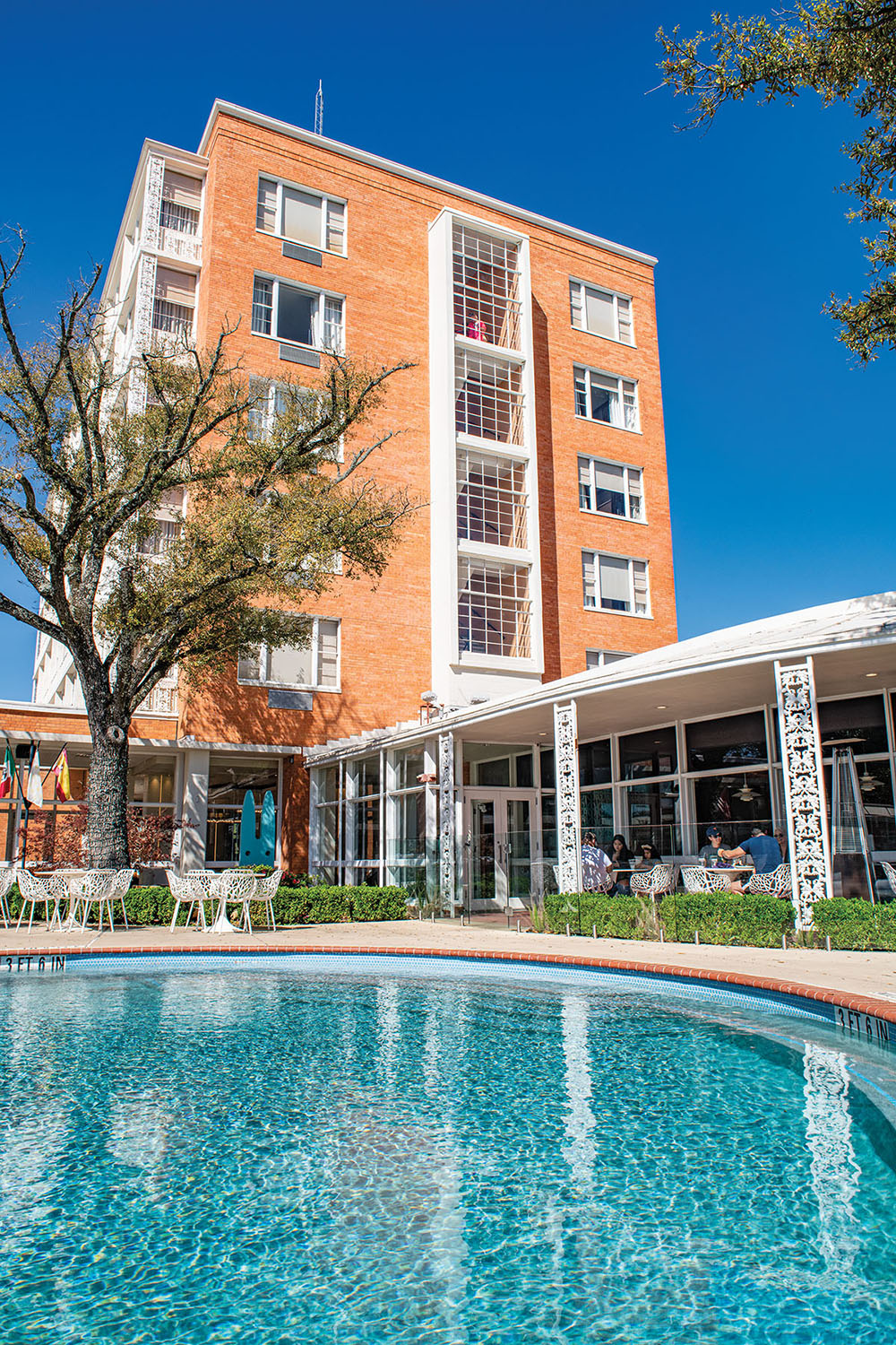 A tall brick hotel stands above a glittering blue pool with white furniture around