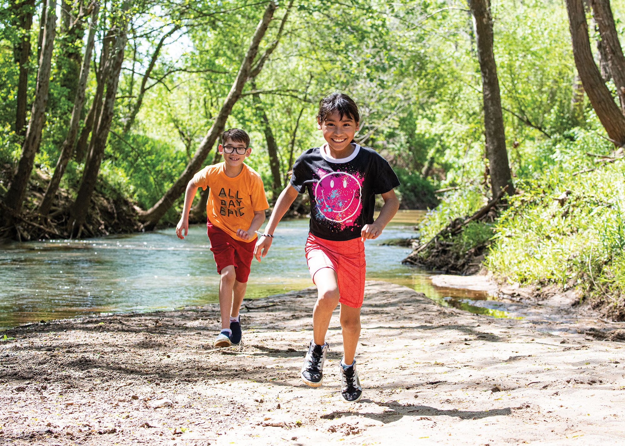 Two young people run along a dirt trail near clear water and green trees