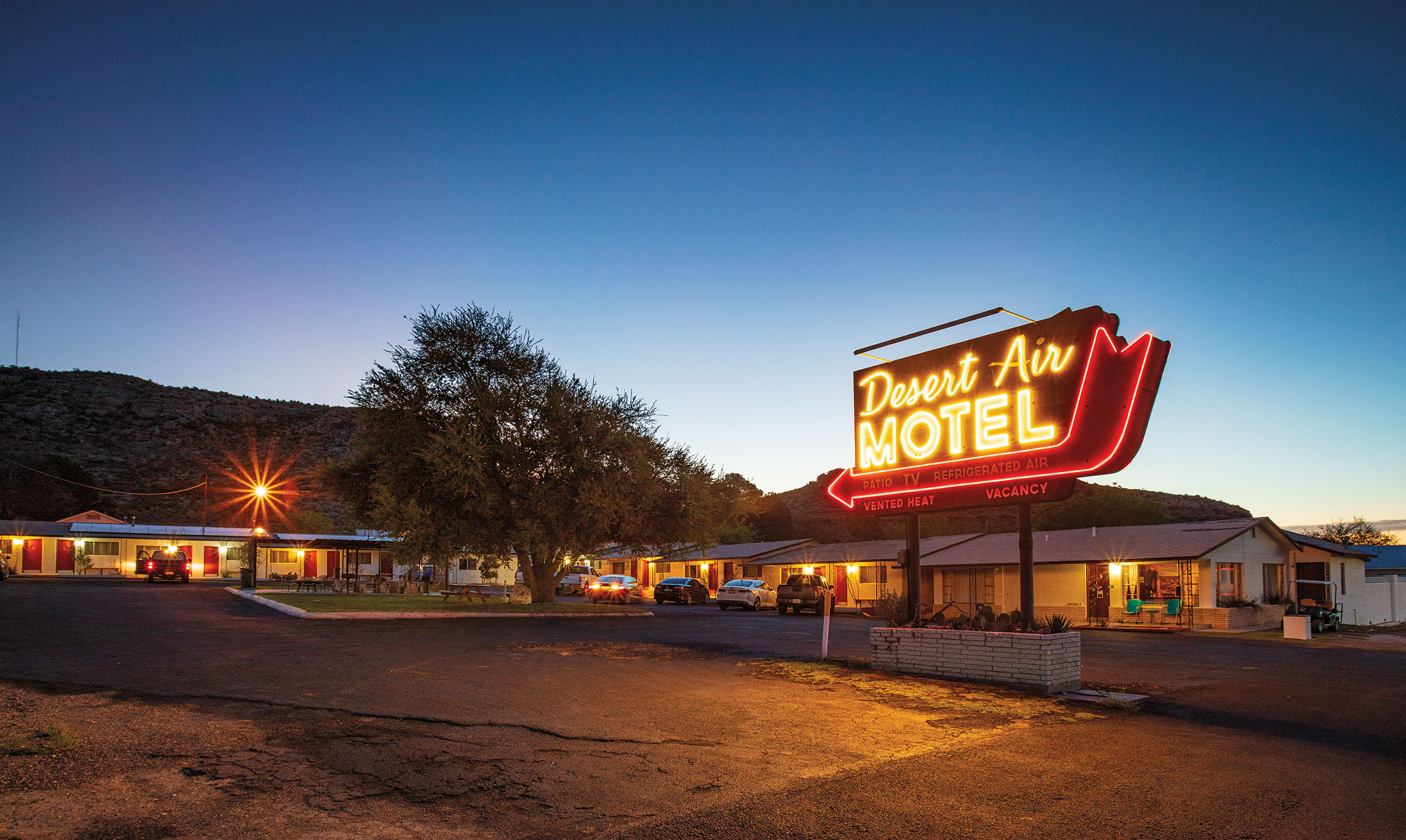 The exterior of a motel with a large neon sign reading 'Desert Air Motel' with cars parked along the front