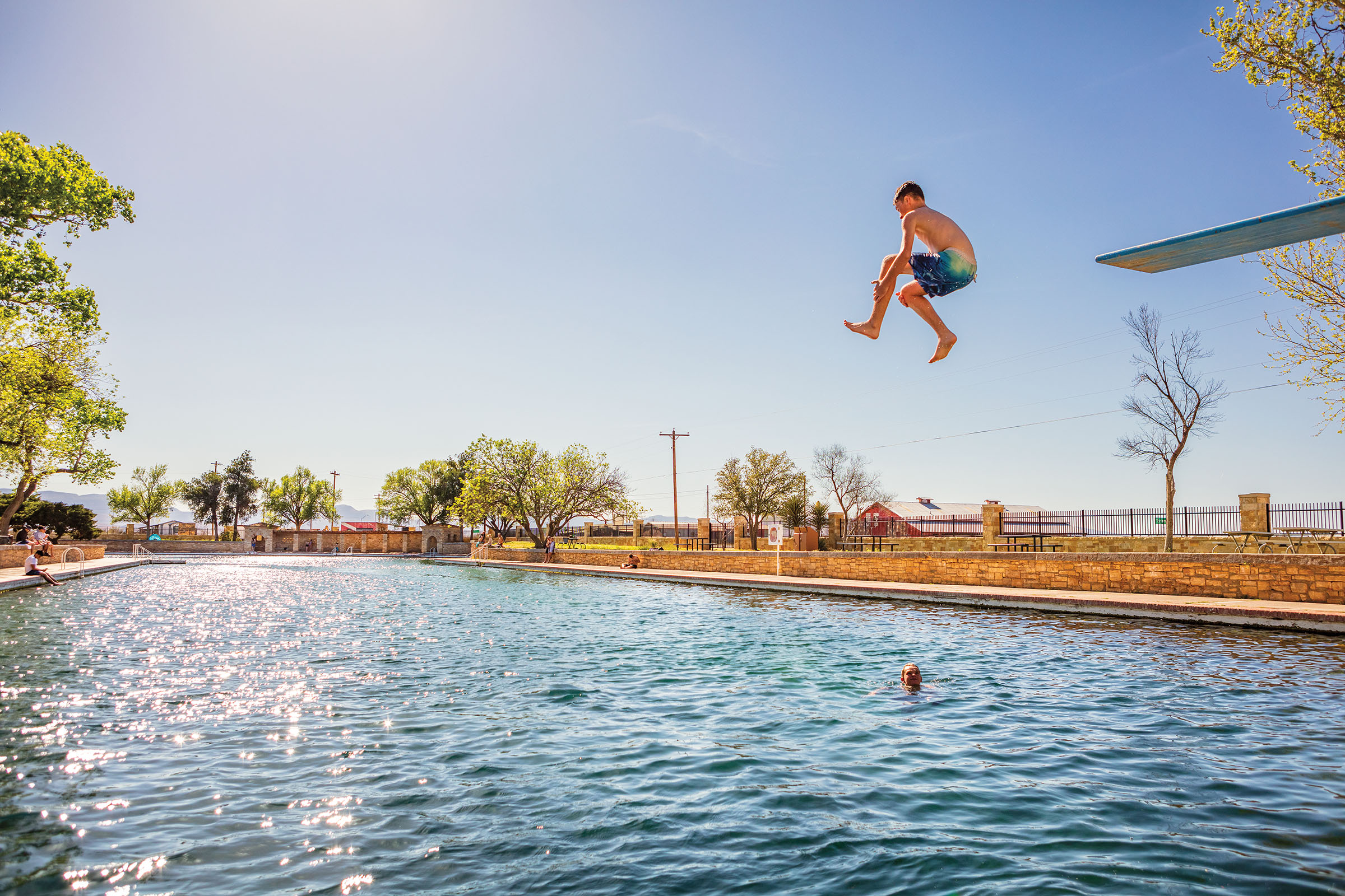 A person jumps off of a diving board into a glittering pool under blue sky