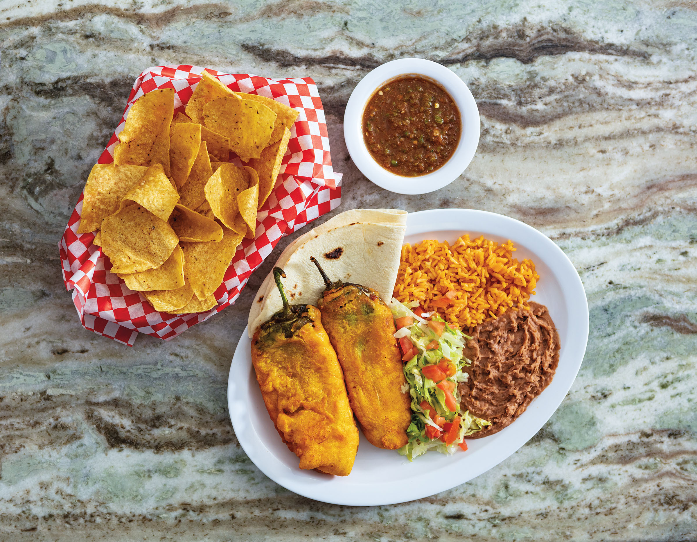 An overhead shot of chile rellenos and rice and beans alongside a basket of golden chips and red salsa