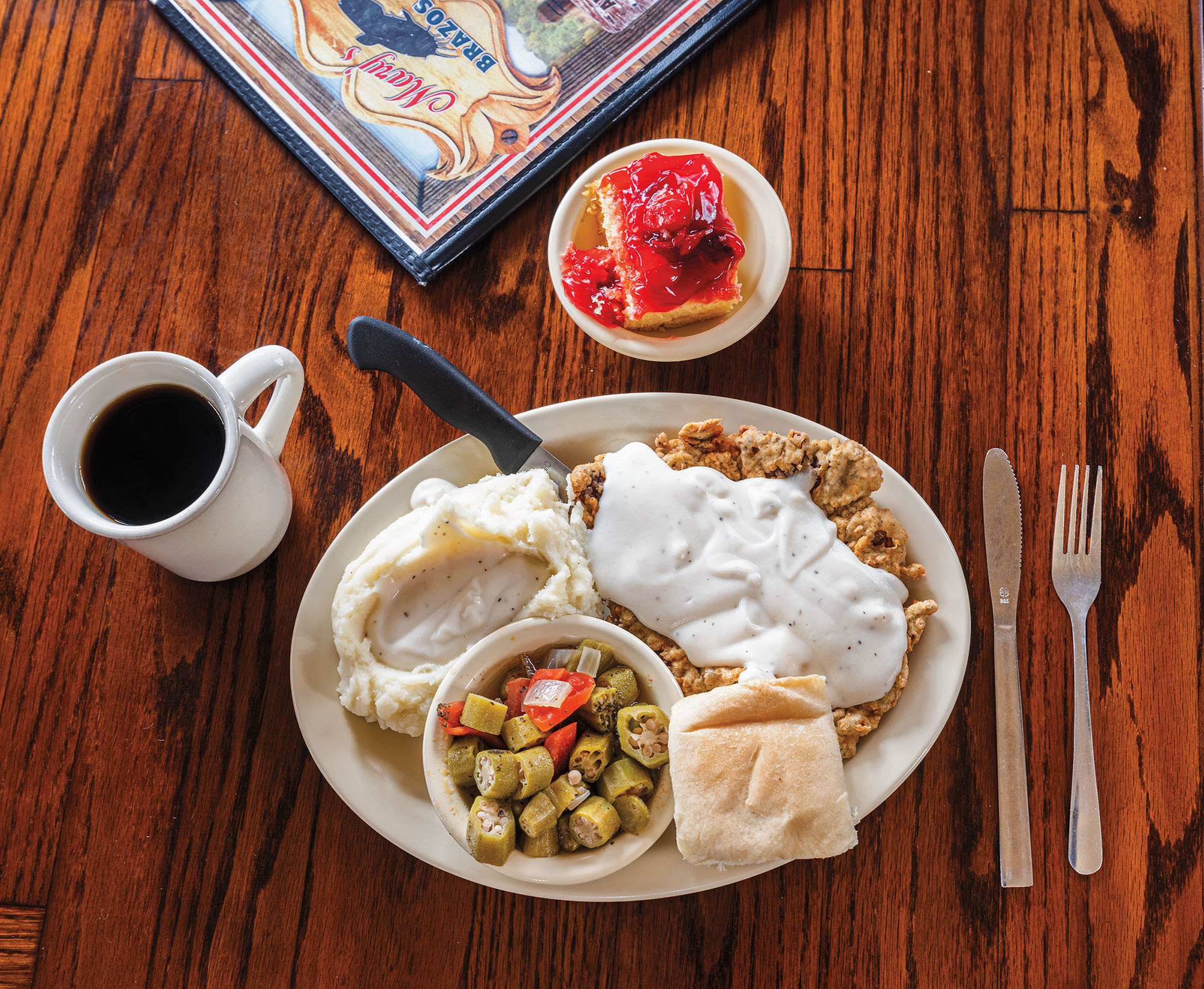 An overhead view of a large chicken fried steak and okra on a white plate on a brown table