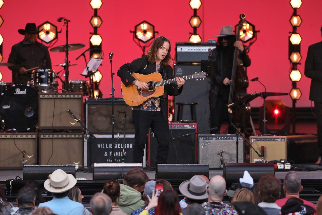 A person holds a guitar on a stage with a red background. An equipment box is lettered with a white stencil reading 