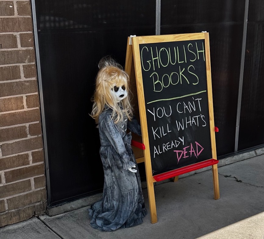 A small creepy doll with ghoulish black outlined eyes and blond hair wearing a raggedy old gown stands next to a chalkboard sign. The greeting on the sign reads: "GHOULISH BOOKS: YOU CAN'T KILL WHAT'S ALREADY DEAD."