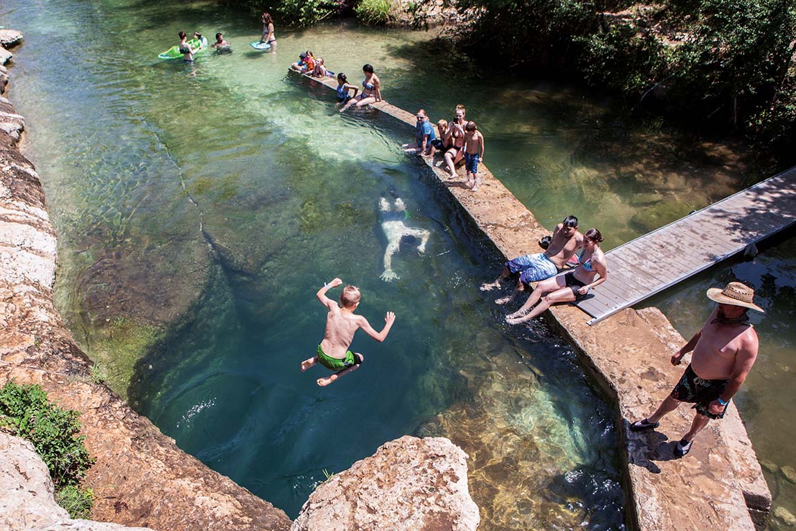 A boy jumps into a deep swimming hole with spectators watching from a nearby dock.