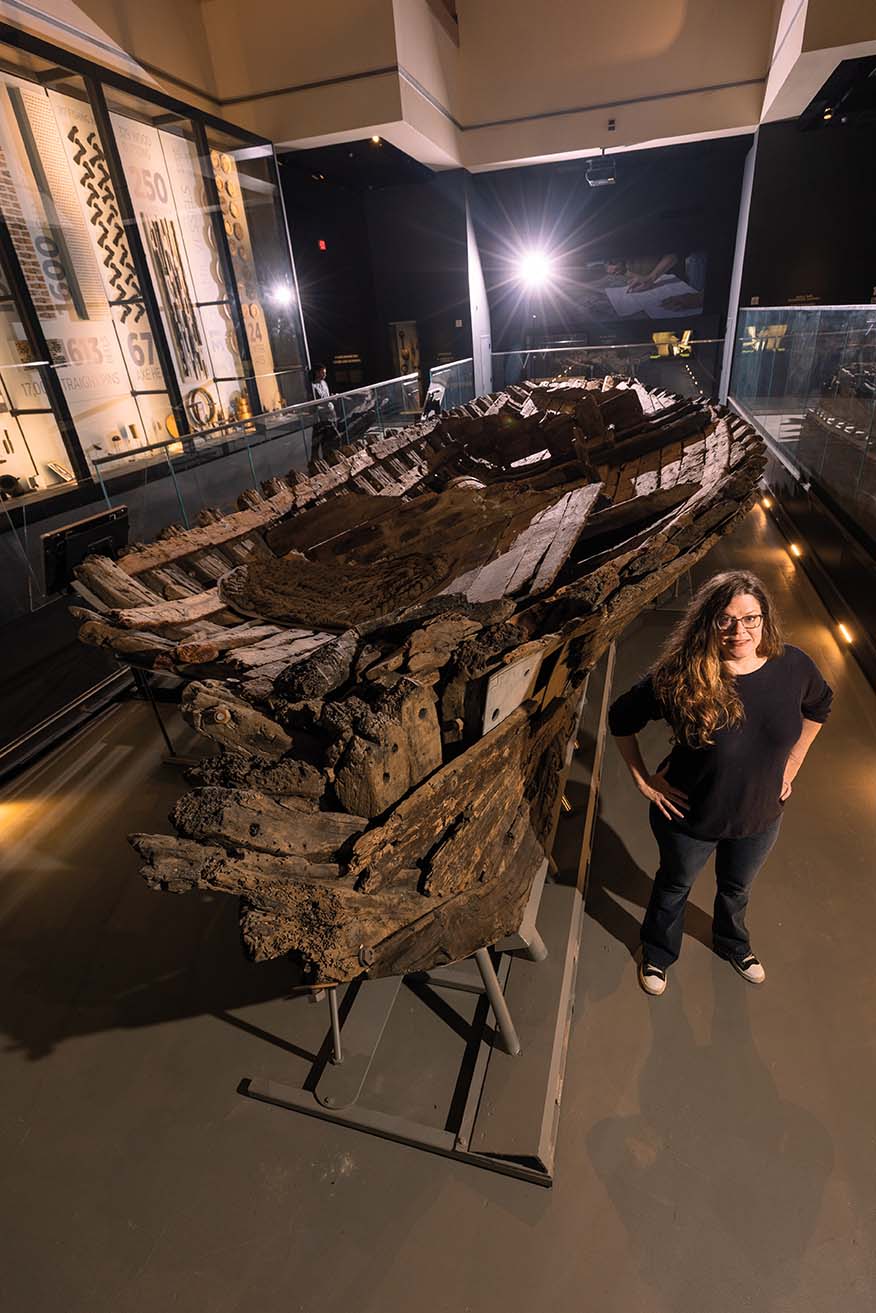 A woman stands proudly in front of a museum display of a shipwreck.
