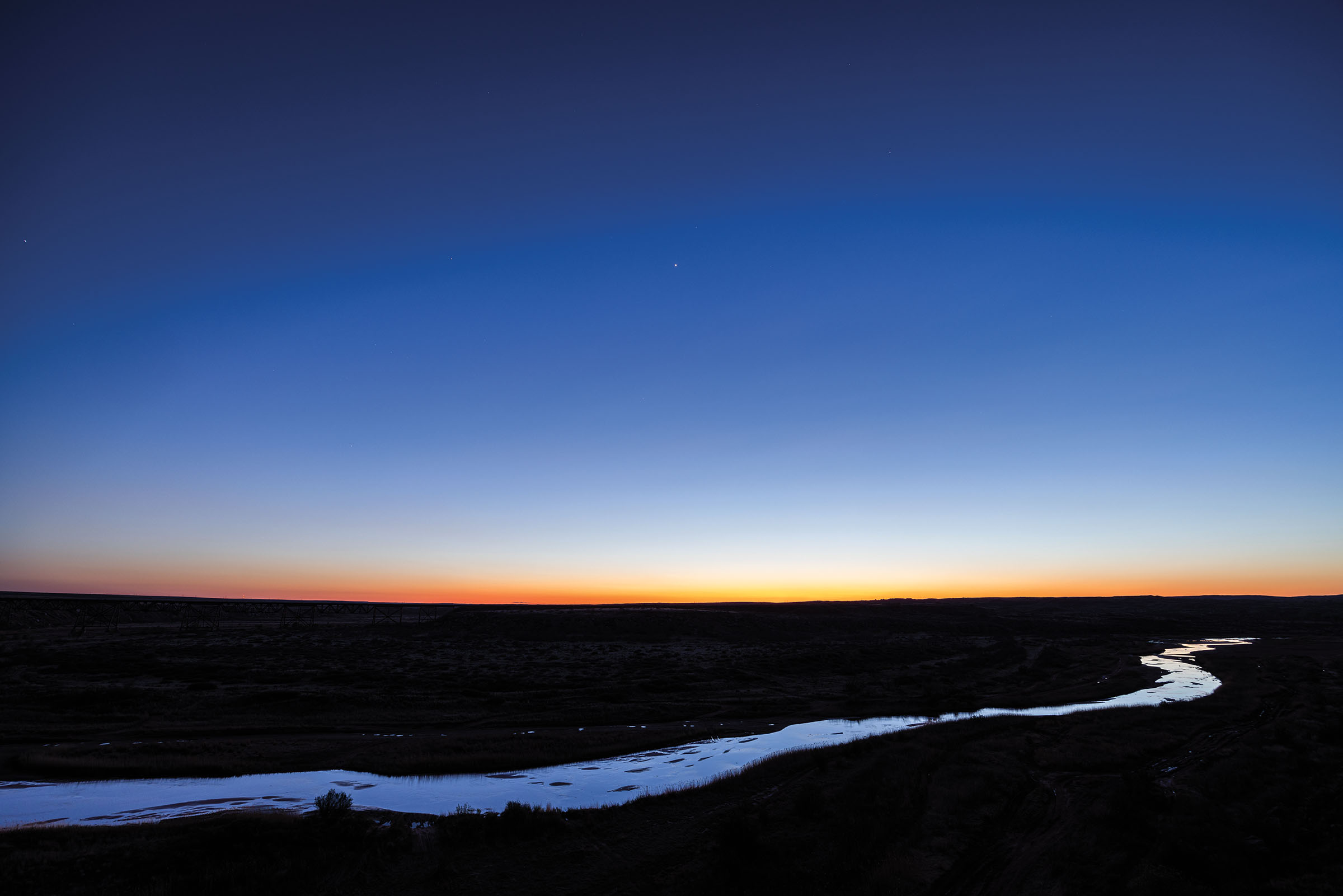 The sun sets turning the sky a deep blue-purple over a strip of blue water, the Canadian River, through the landscape