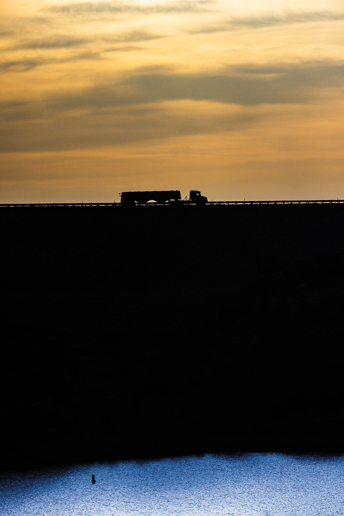 The silhouette of a truck passing along a bridge above dark blue water