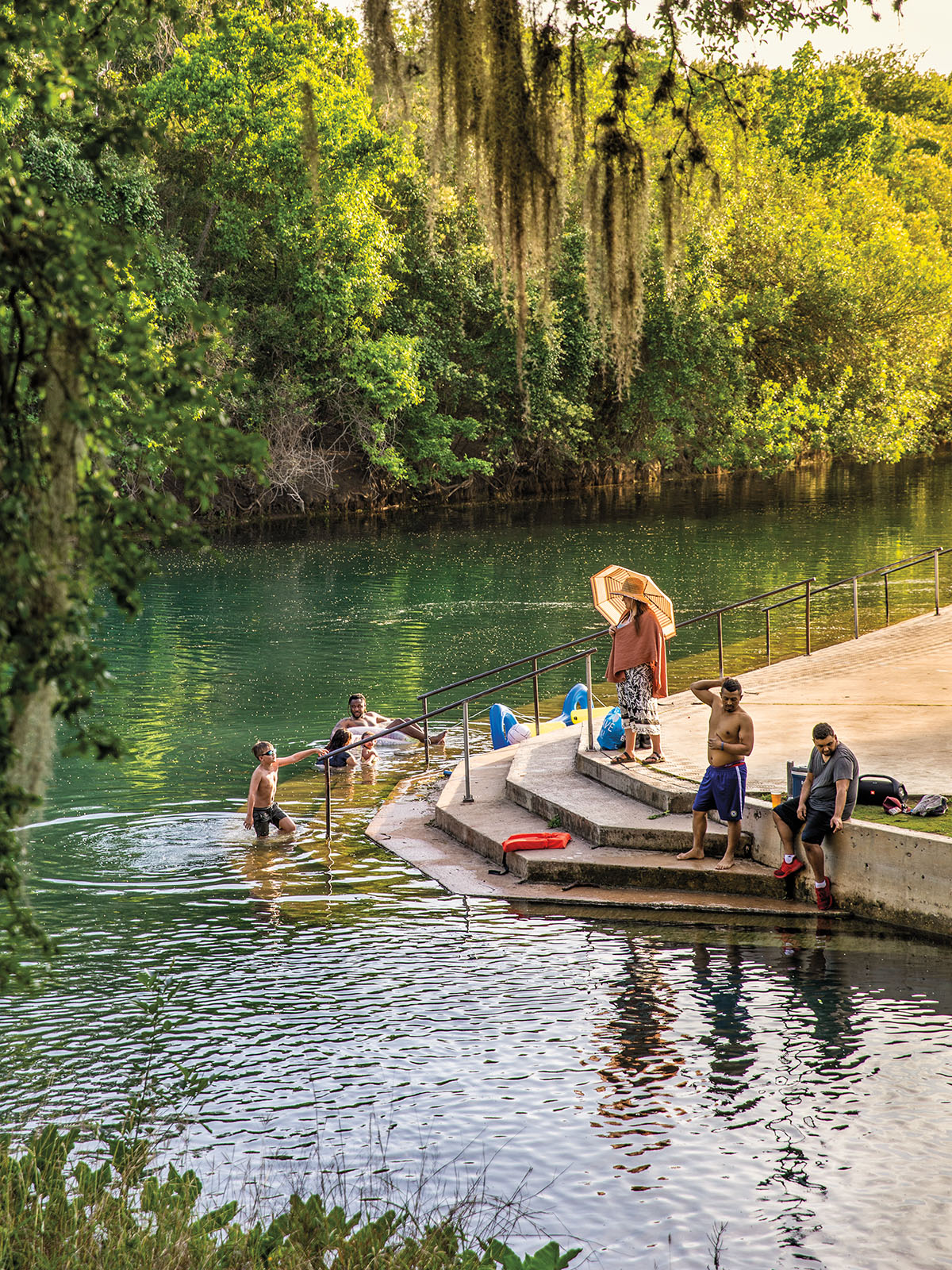 A small group of people sit on concrete steps and swim in a river scene under green trees