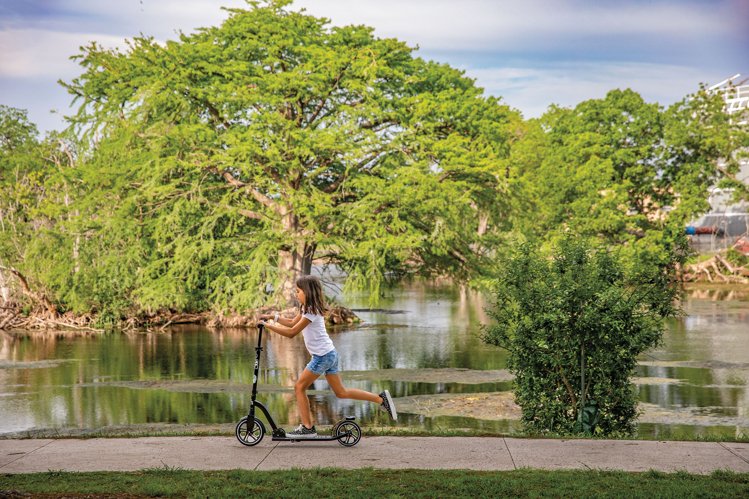 A young woman rides a scooter on a concrete sidewalk in front of tall green trees and a riverfront