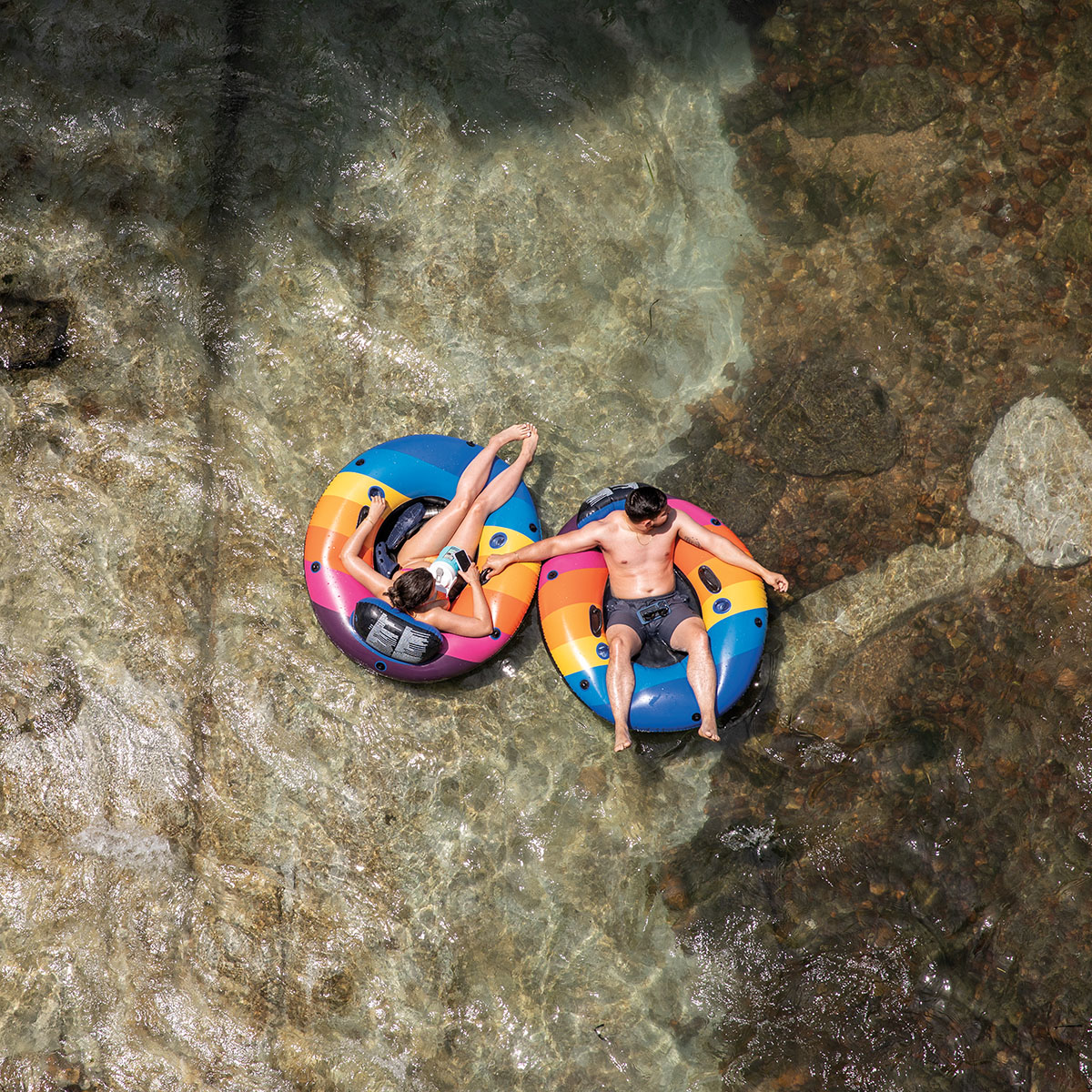 An overhead view of two people relaxing and holding hands in brightly colored inflatable tubes on clear water