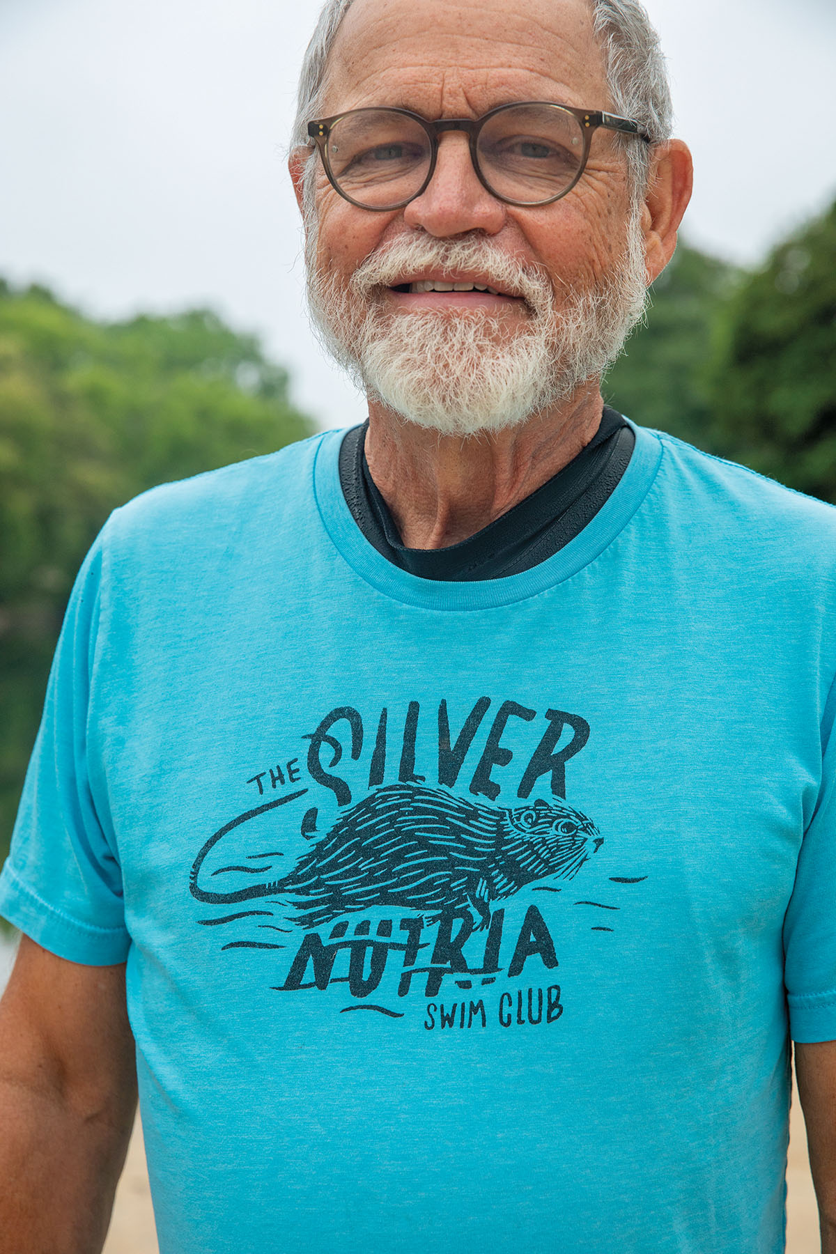 A man with a short white beard wears a teal shirt reading "Silver Nutria Swim Club"