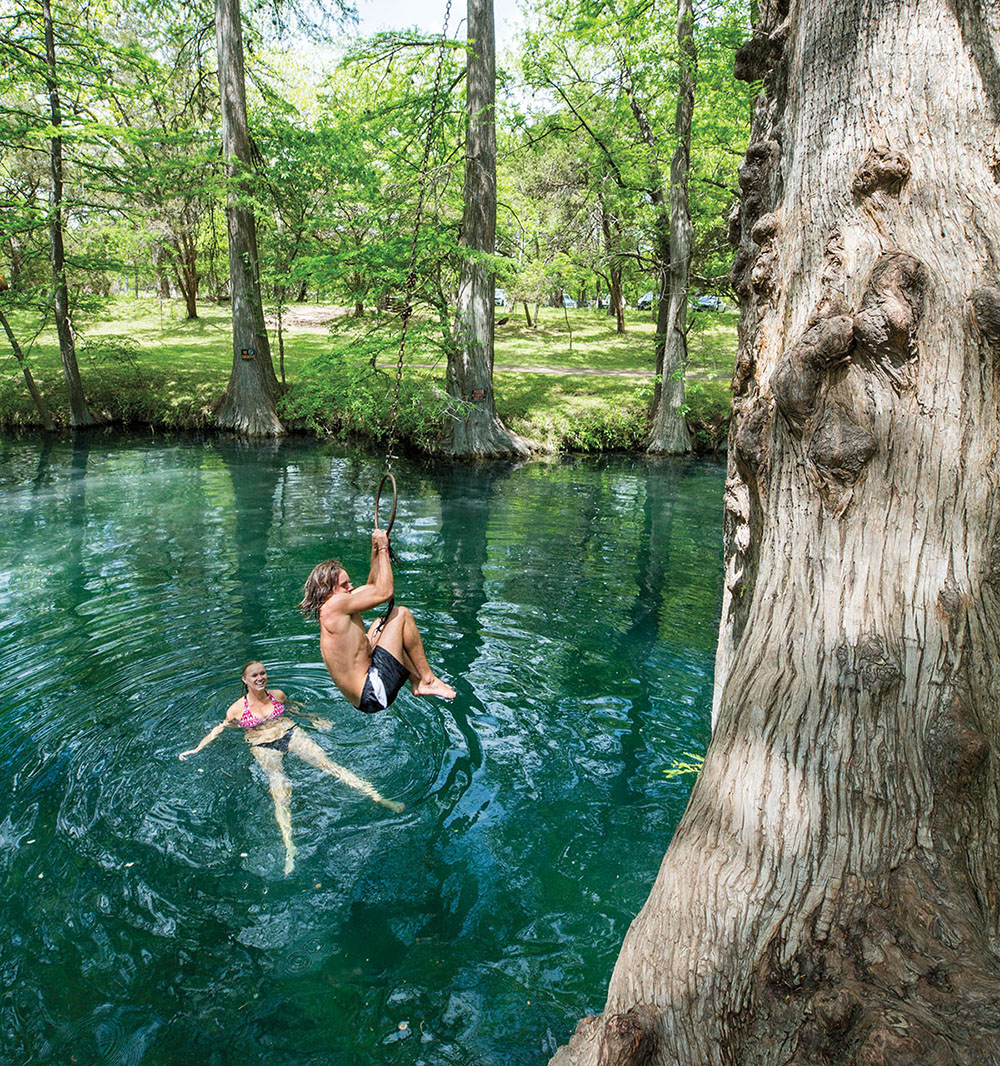 One person swims while another swings from a rope swing into beautifully clear blue green water