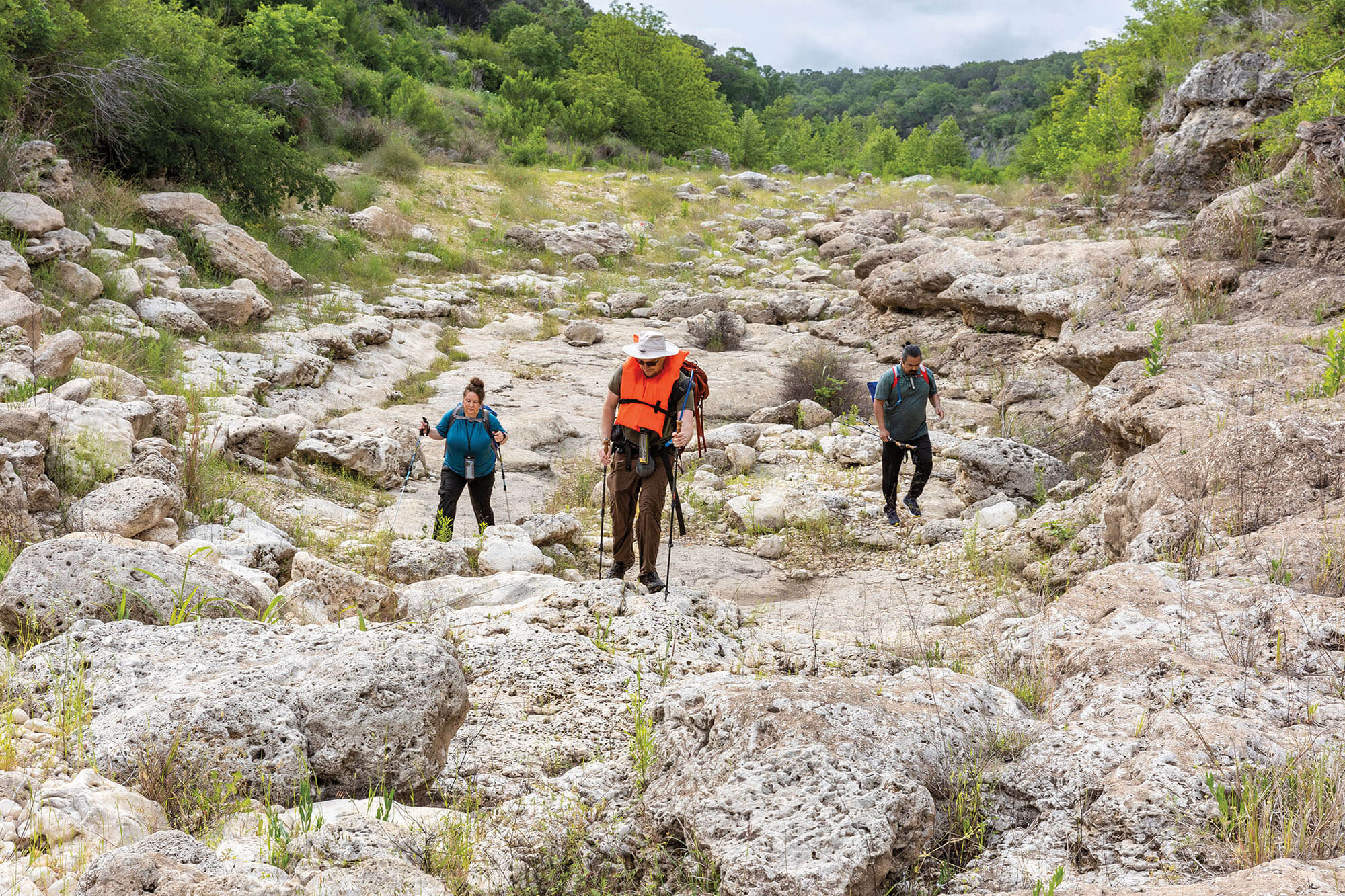 A group of people, one wearing an orange life vest, traverses a rocky landscape under green trees