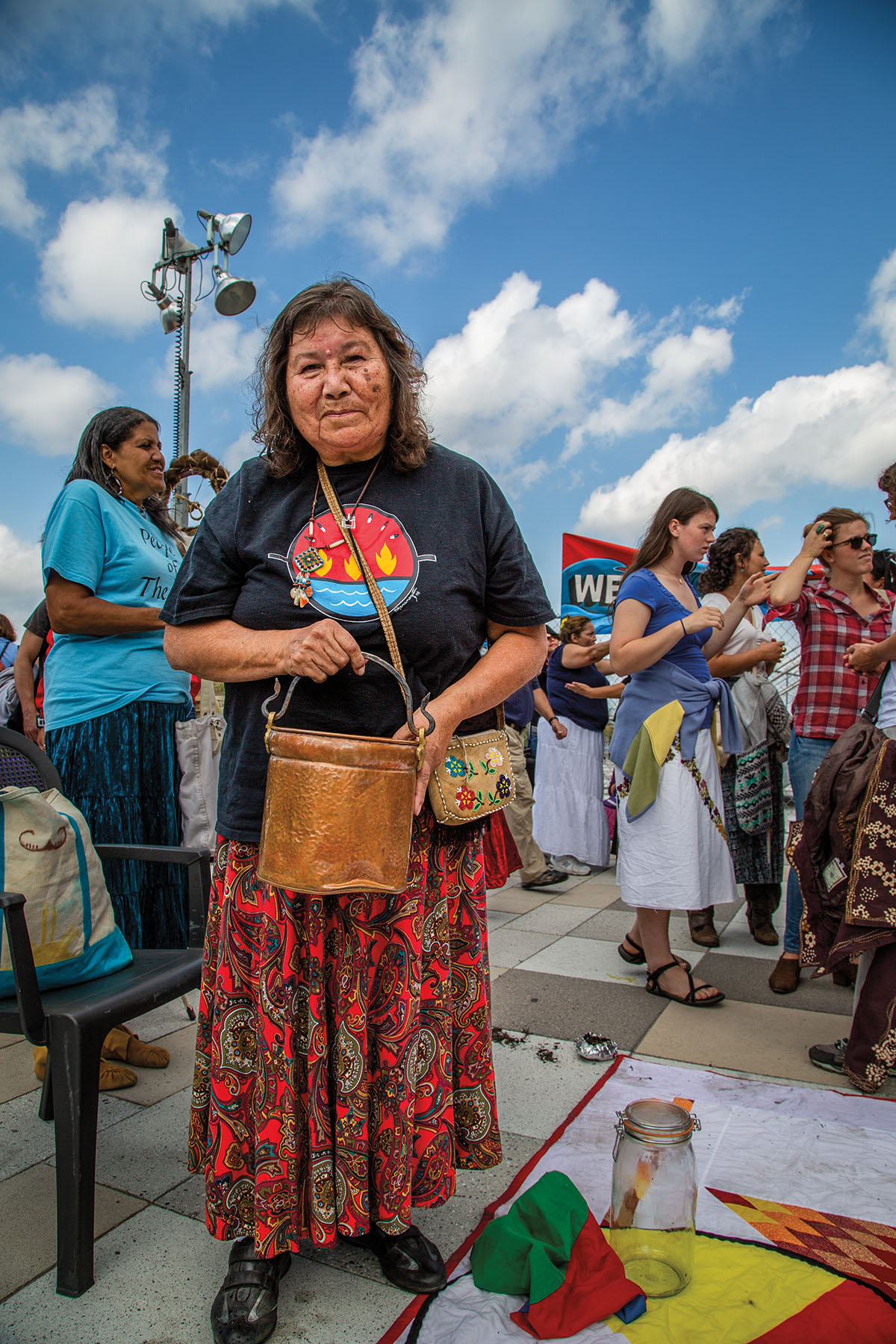 A person in a long red skirt and gray t-shirt holds a copper bucket under blue sky